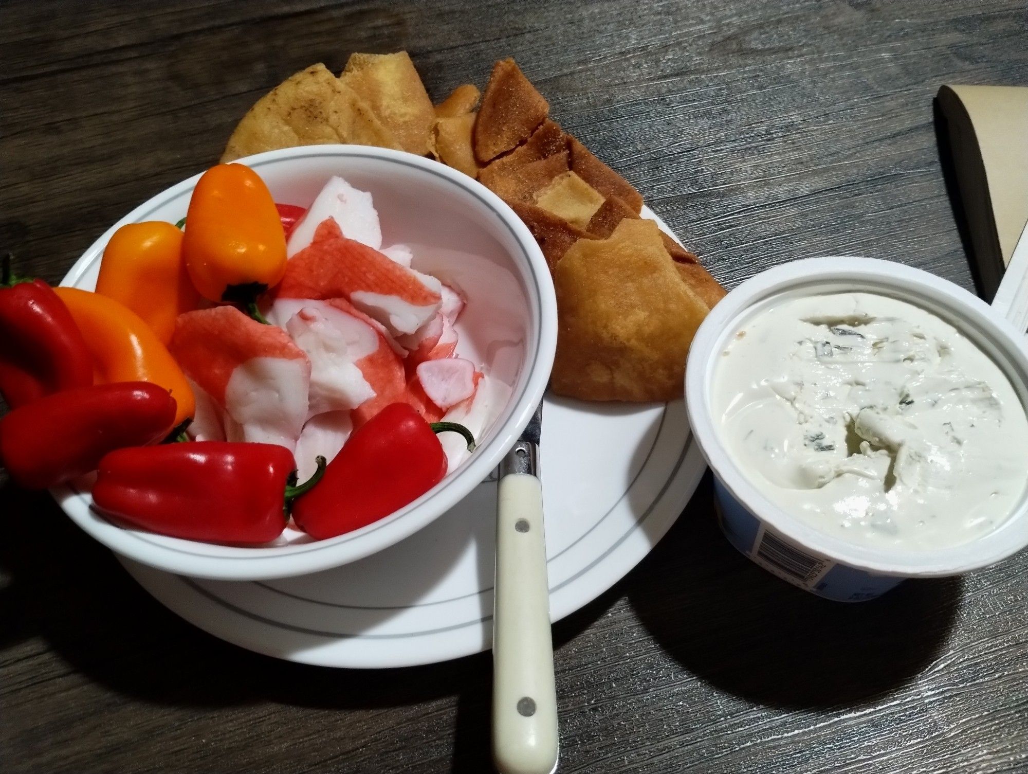 A tub to the right with chive and onion cream cheese. A white bowl sitting atop a white plate with a white handled butter knife. In the bowl is surumi and mini sweet peppers. On the top part of the plate is pita chips. The surface is a dark wood print, wood textured desk top.