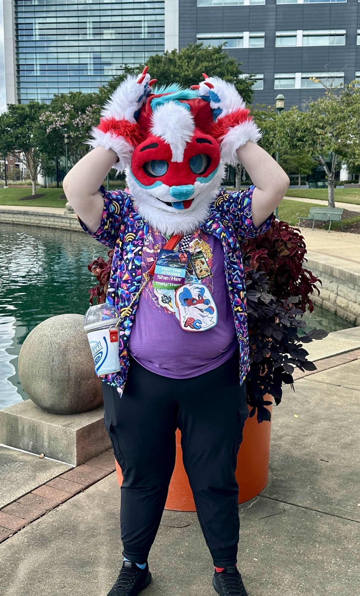 A red/blue/white dog partial fursuiter in a purple outfit holds their floppy ears above their head. They are standing in front of a planter near a man-made lake in a city landscape.