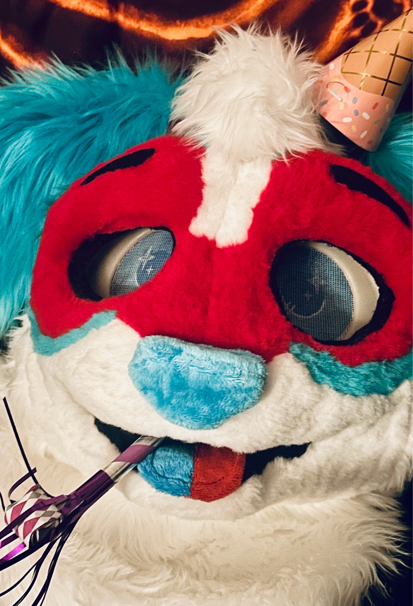 Selfie of a red/blue/white dog fursuiter with a party hat and party horn in their mouth