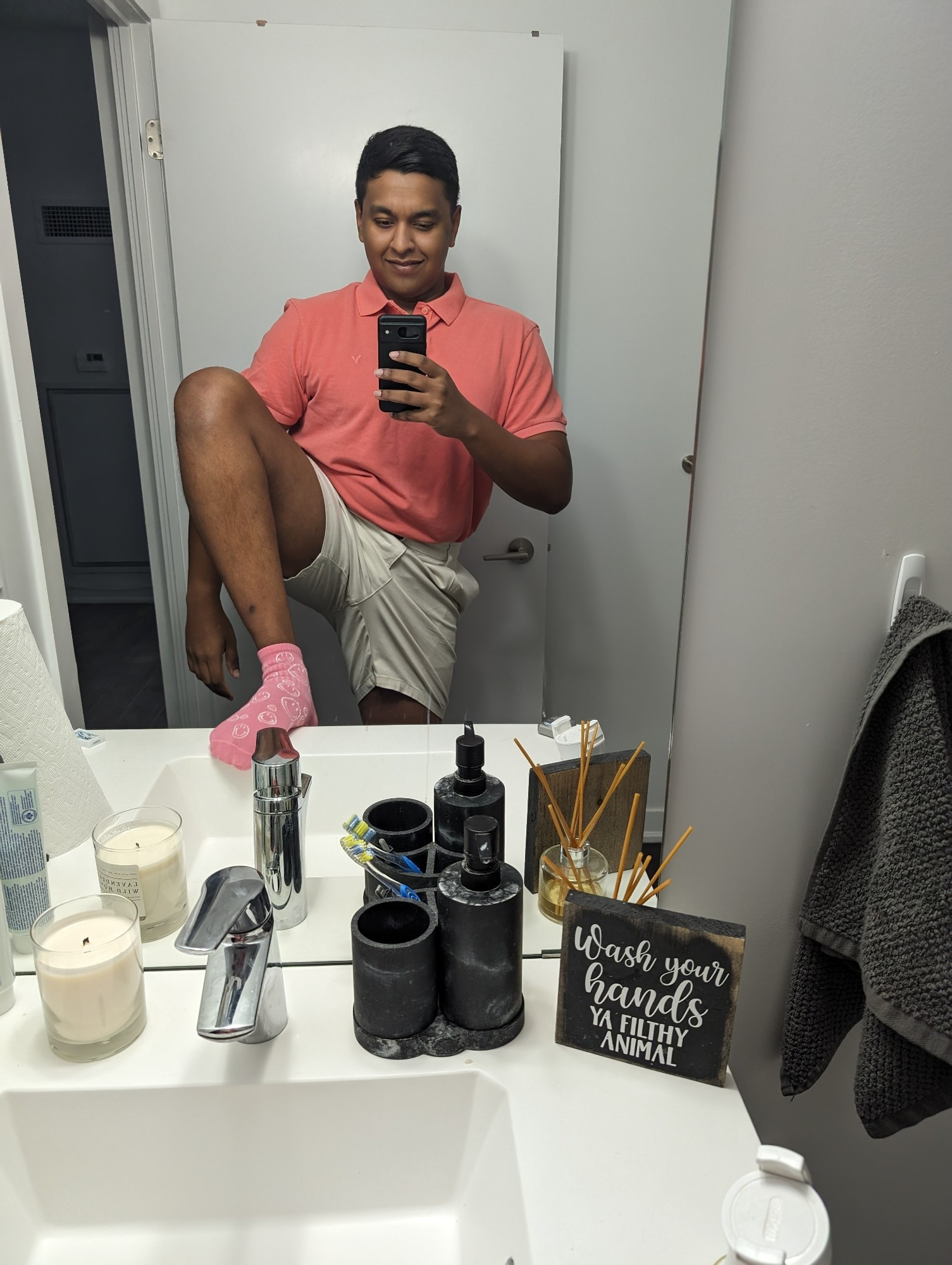 Bathroom mirror selfie of a brown skinned 5'10" man smiling in a salmon pink polo shirt tucked into white shorts. One foot rests on the bathroom sink, showcasing his cute pink socks with smiley faces on them. He is barbiecore and barbiecore is he. 