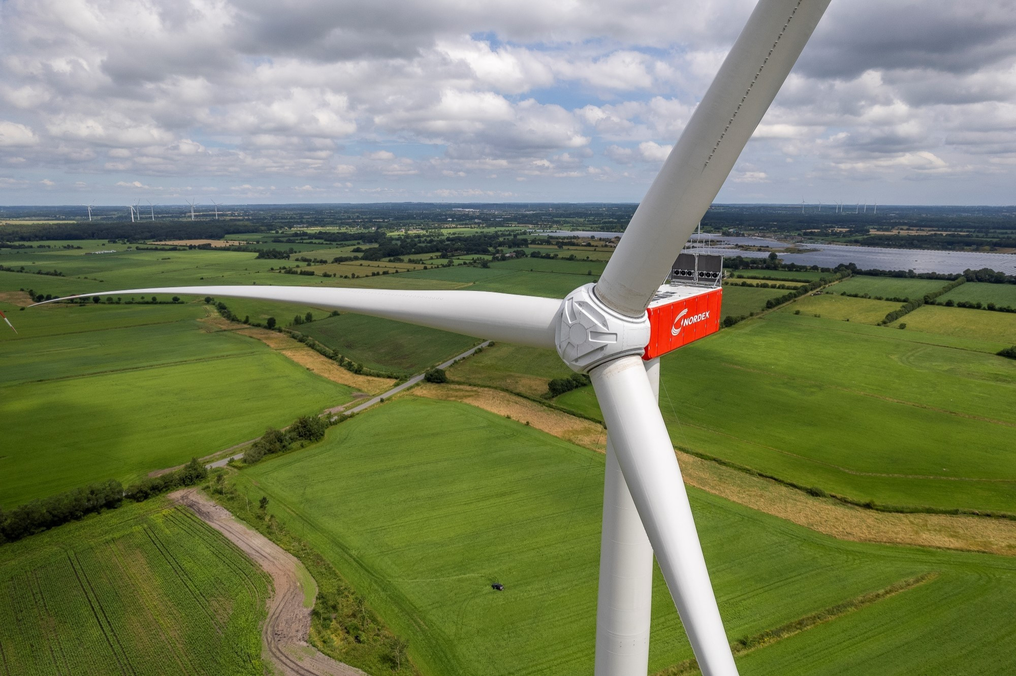 Große Windenergieanlage Nordex N175 vor grüner offener Kulturlandschaft mit Gewässer im Hintergrund und bewölktem Himmel. Das Foto ist etwa auf Nabenhöhe der Anlage (Mittelpunkt des Rotors) aufgenommen.