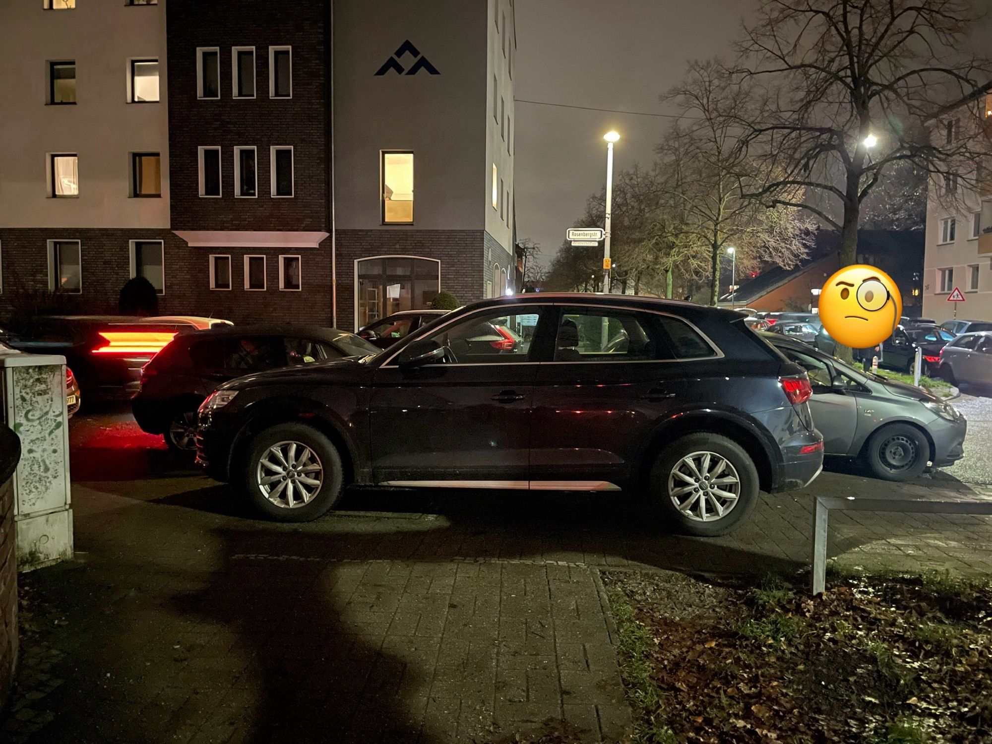 Ein Audi SUV wurde auf dem Gehweg abgestellt. Wer von dem querenden Gehweg kommt, muss über eine Grünfläche mit niedriger Metalleinzäunung ausweichen.