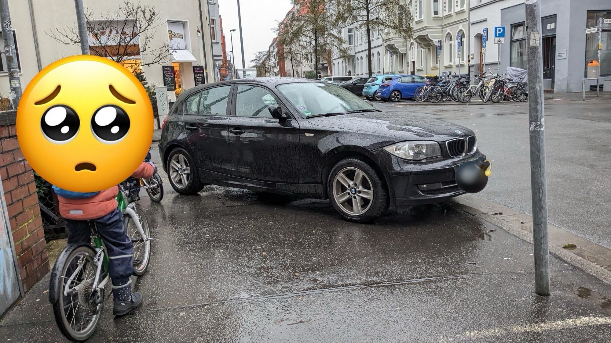 Blick vom Gehweg aus auf eine Straßenkreuzung. Zwei kleine Kinder sind mit ihren Fahrrädern auf dem Gehweg unterwegs. 
Ein schwarzer BMW wurde im Einmündungsbereich der Kreuzung quer auf dem Gehweg abgestellt und blockiert die sichere Querung der Kinder.