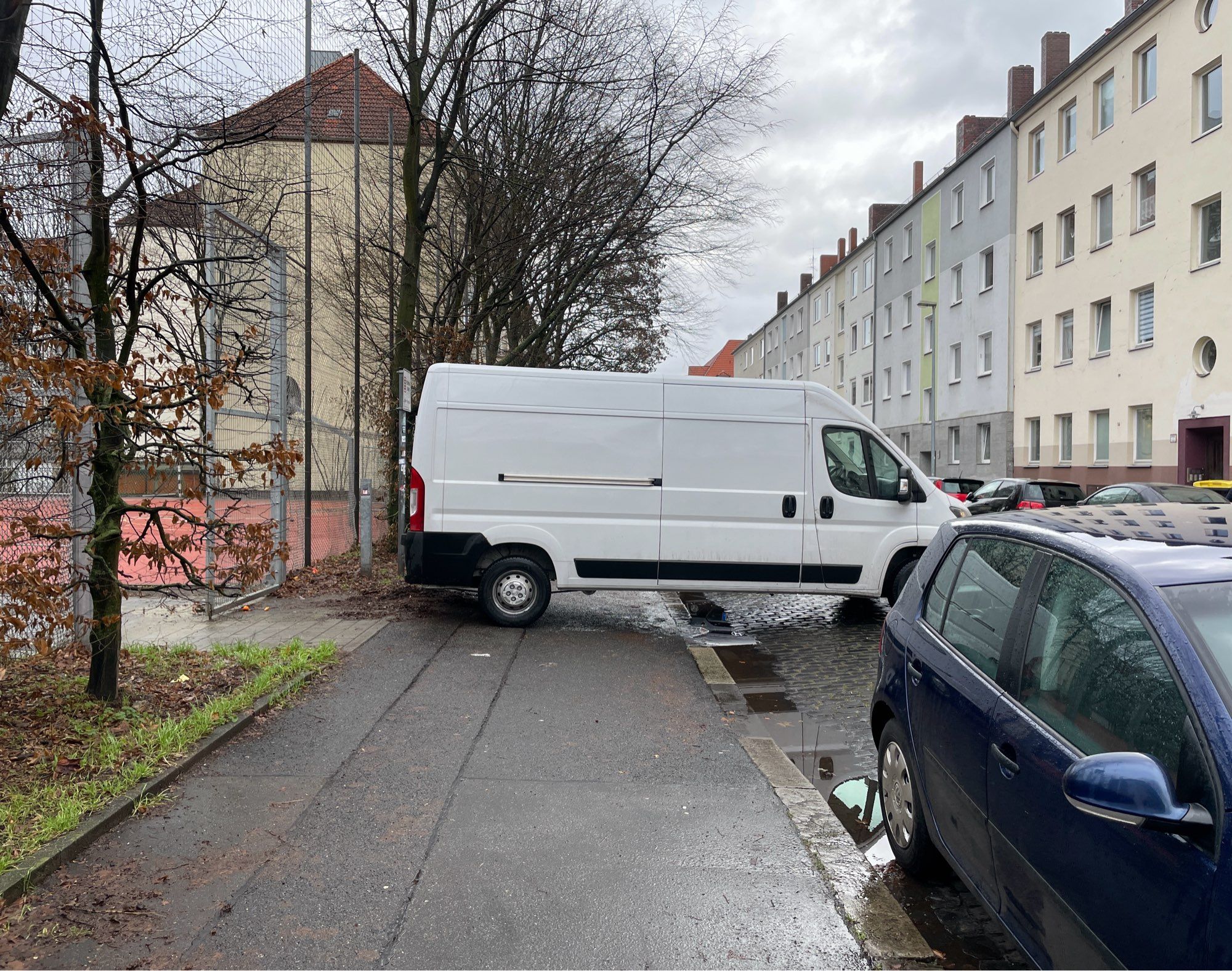 Blick entlang eines Gehwegs. Ein weißer Transporter wurde quer über selbigen abgestellt und blockiert den Weg.