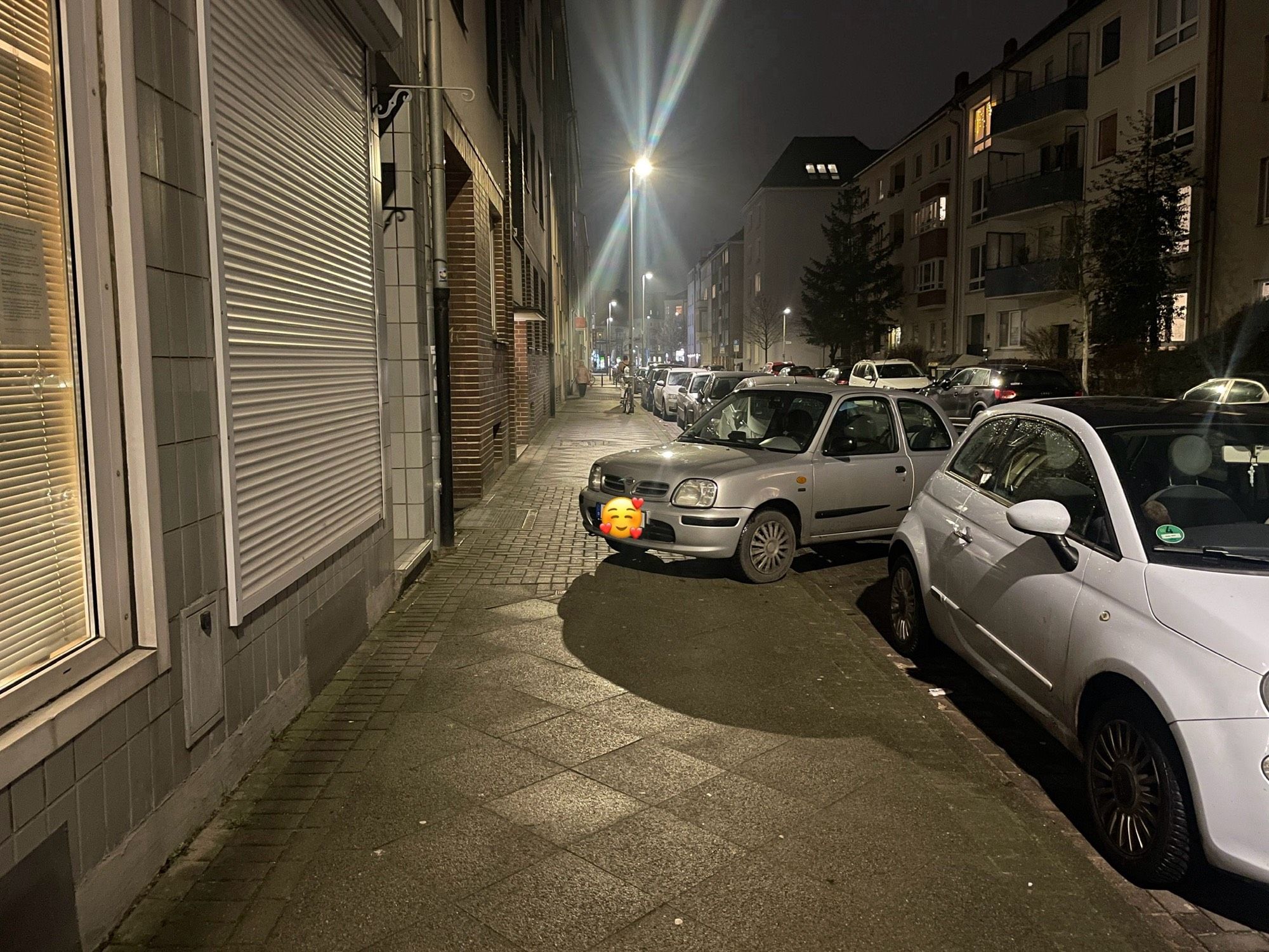 Blick entlang eines Gehwegs. Ein silberner Kleinwagen wurde quer zur Fahrbahn geparkt und blockiert dadurch den Großteil des Gehwegs