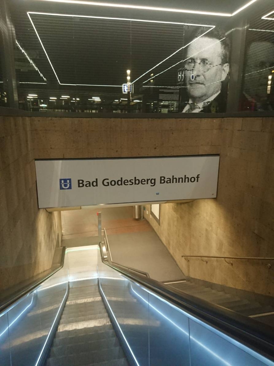 Photo of the entry of metro station "Bad Godesberg Bahnhof" in Bonn with the glass ceiling in the upper half of the picture and stairs and the escalator in the lower half. Through the paintet window with the image of Paul Löbe, the metro sign and streetlights can be seen. The neon lights of the stairs and the ceiling reflect in the glasses, giving a futuristic impression.