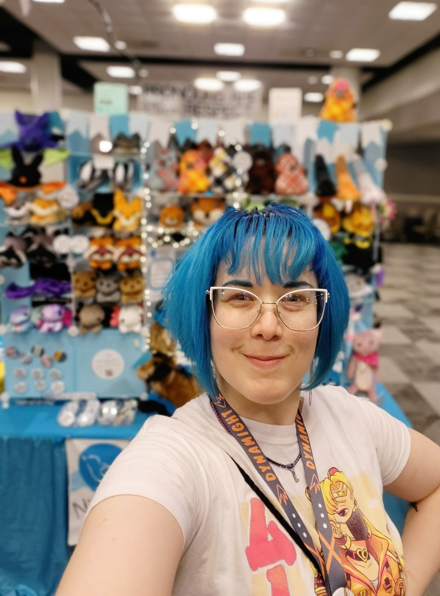 me posing in front of my booth full of plushies and sample plush demonstrating our commissionable styles! there's a lot of blue and cute pennants at the top, and twinkle lights. I'm smiling like I don't feel awkward taking selfies but I might be lying 🤭