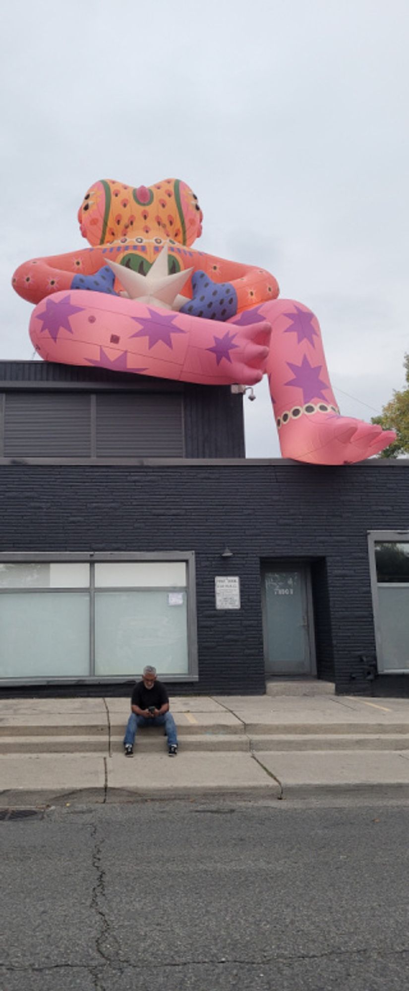 Ventura by Amanda Lobos: a large, colourful, inflatable Janus-faced creature seated atop a large industrial building. Below, a man seated and checking his phone