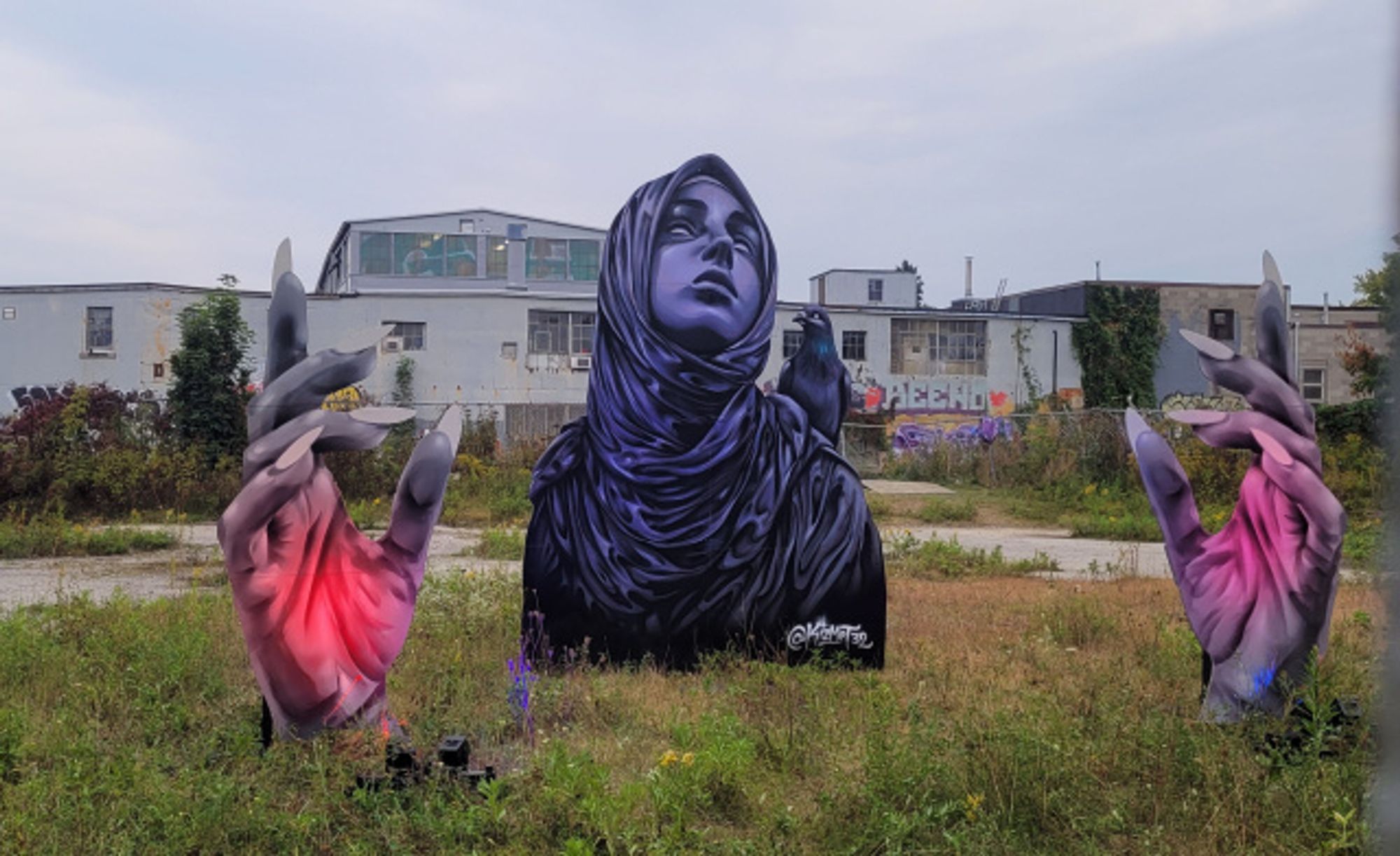 Couldn't find the artist for this one on the website :( A three-part installation of paintings in an industrial lot. The central painting is the bust of a woman wearing a hijab looking upward with a pigeon on her shoulder. On either side are paintings of open hands with red light shining onto each palm