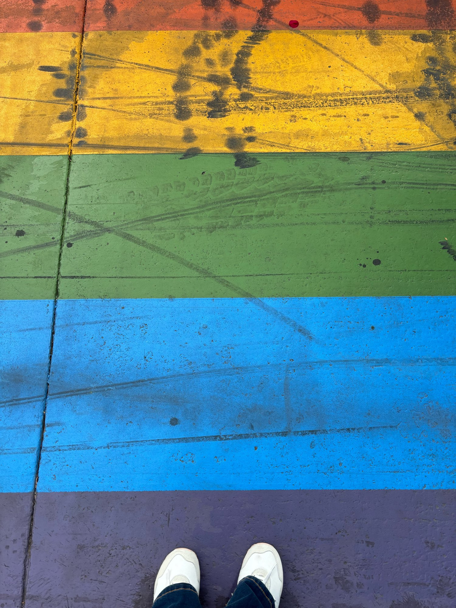 A pair of white sneakers stick out onto a rainbow striped walking path. The colors are shown horizontally: orange, yellow, green, blue, and purple. A series of tire marks are strewn across the path.