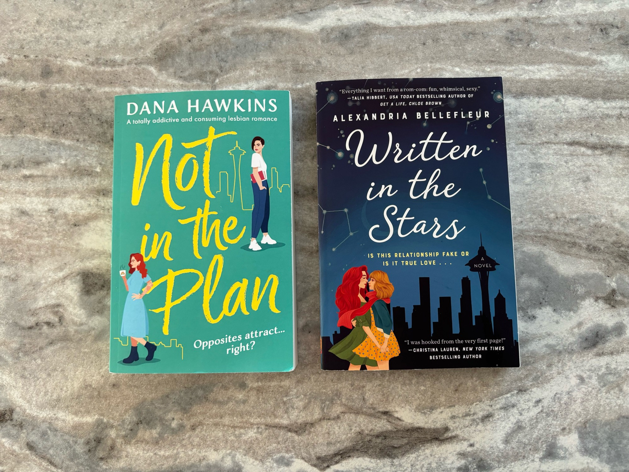 Two books lay on a gray marbled counter. The one on the left has a green cover with yellow text, and two women set against an outline of Seattle. Its title is “Not in the Plan” by Dana Hawkins. The second book has a night sky cover with “Written in the Stars” by Alexandria Bellefleur in curvy white font. It has a dark silhouette of Seattle, and also features two women. Both books seem really, really gay.