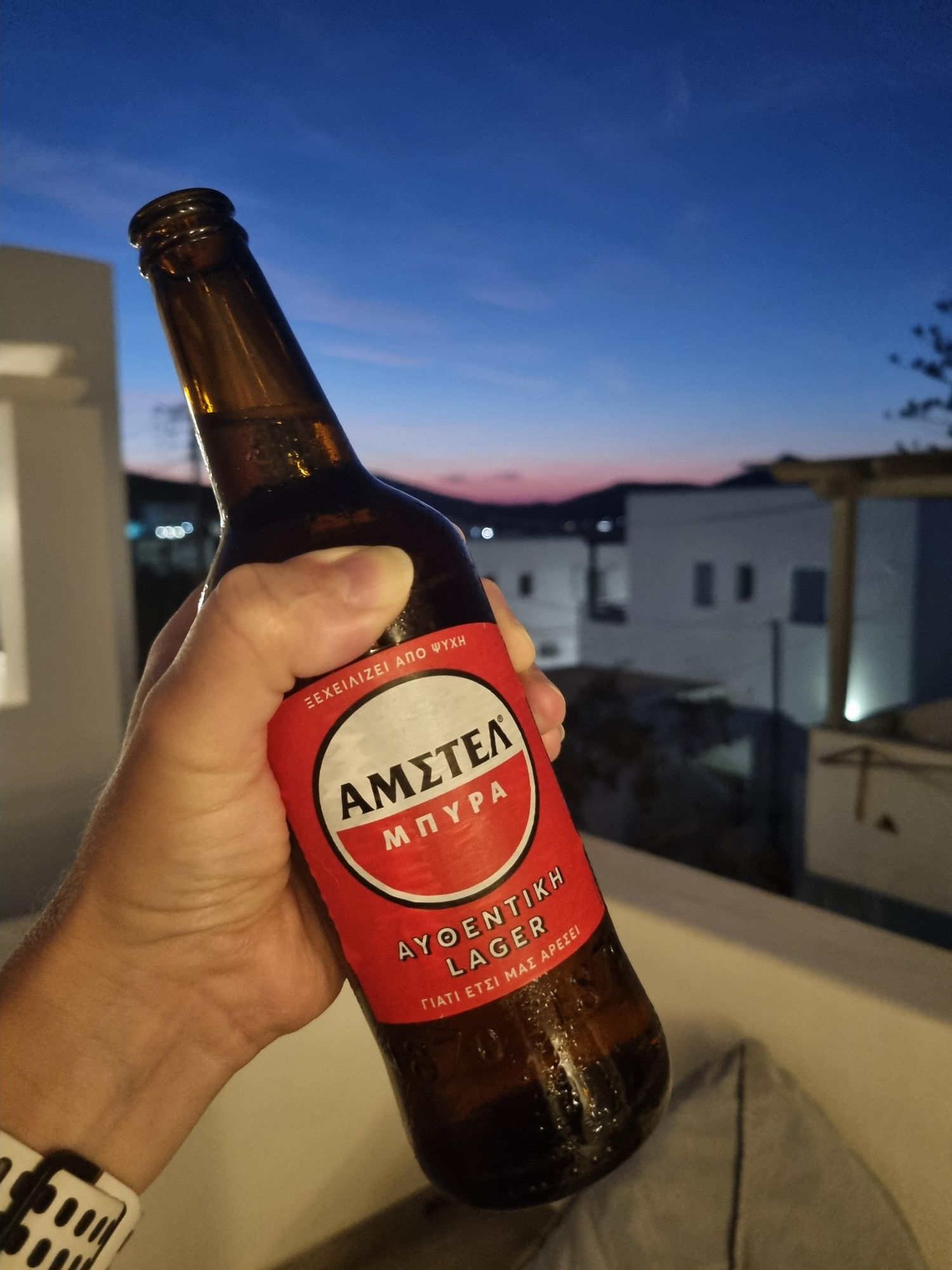 i'm holding a bottle of Greek Amstel Lager. In the background there are some little white houses, far away a part of a Greek harbour town and the sun going down behind some rocks. 