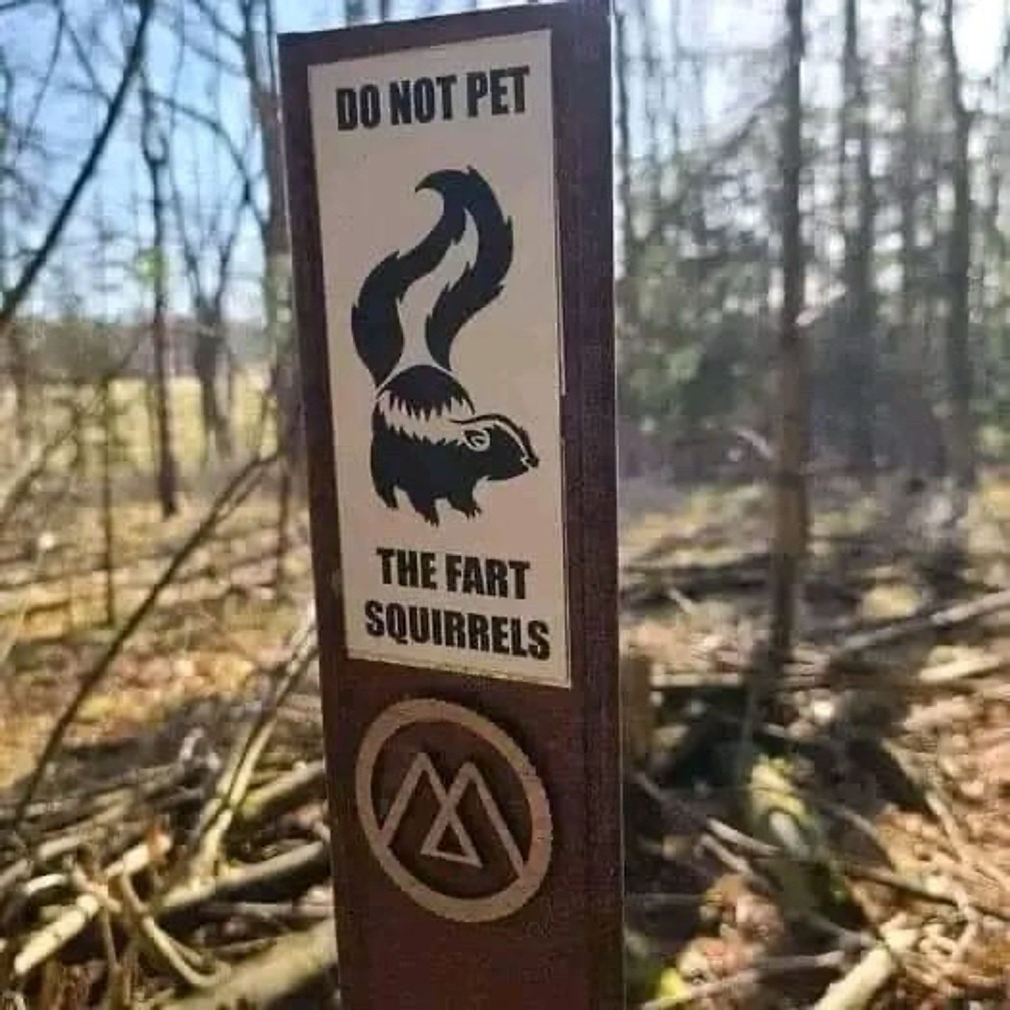 A warning sign with a skunk vertex art that says Do Not Pet The Fart Squirrels in a wooded area with cut down trees