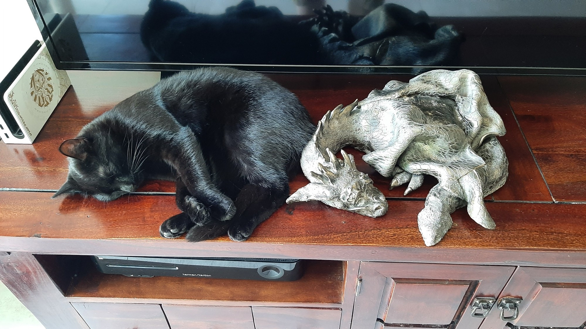 A black cat lies sleeping on it's side, on top of a tv stand, next to a similarly sized statue of a dragon, which lies sleeping in the same position as the cat.