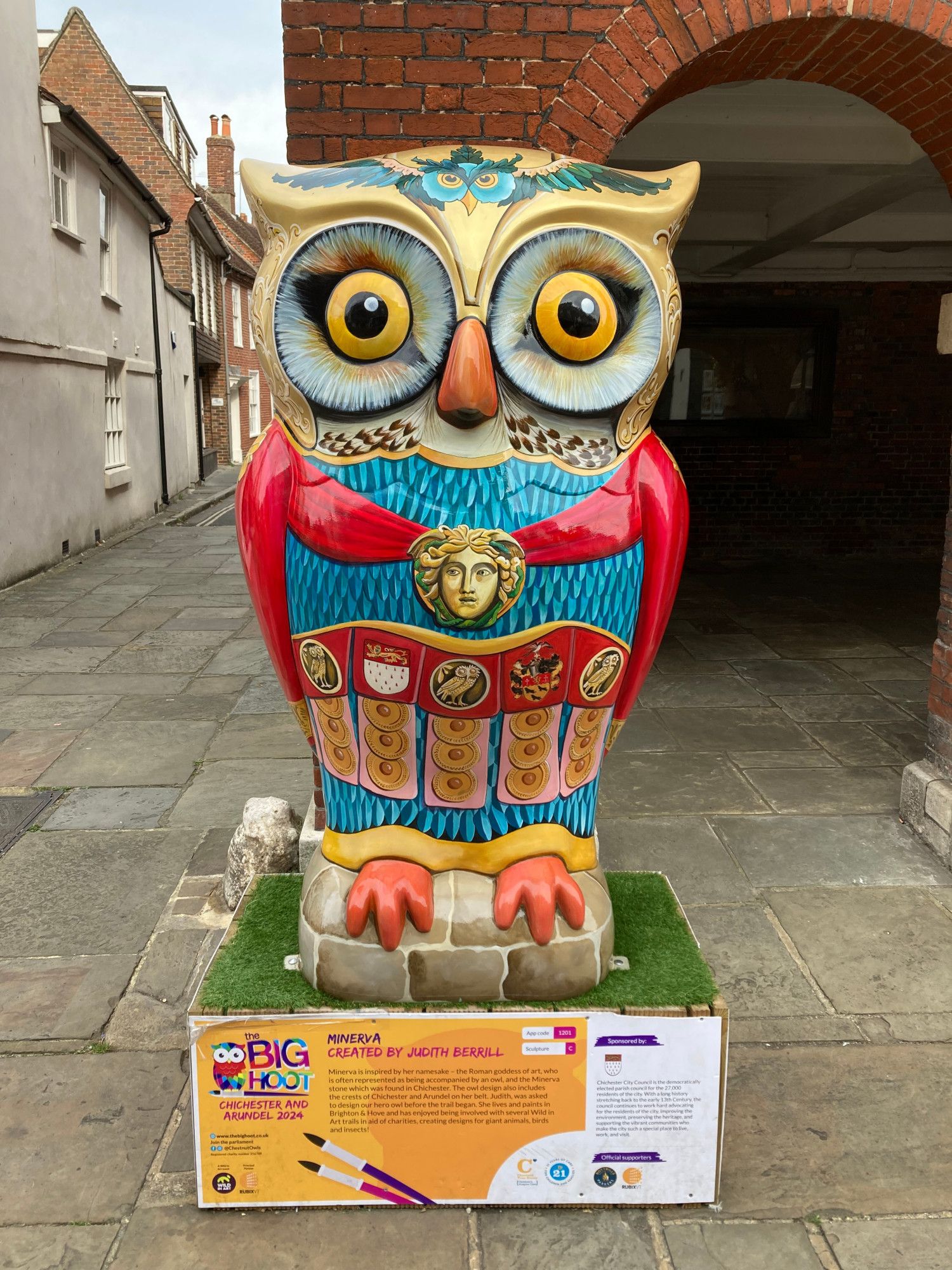 Large owl sculpture with regal painting, red robes painted on and a gold broach in middle of chest and red sash across the middle. An owl with wings is painted across its head and the main colours are blue, red, organge and yellow. The owl sits on a plinth on pavement