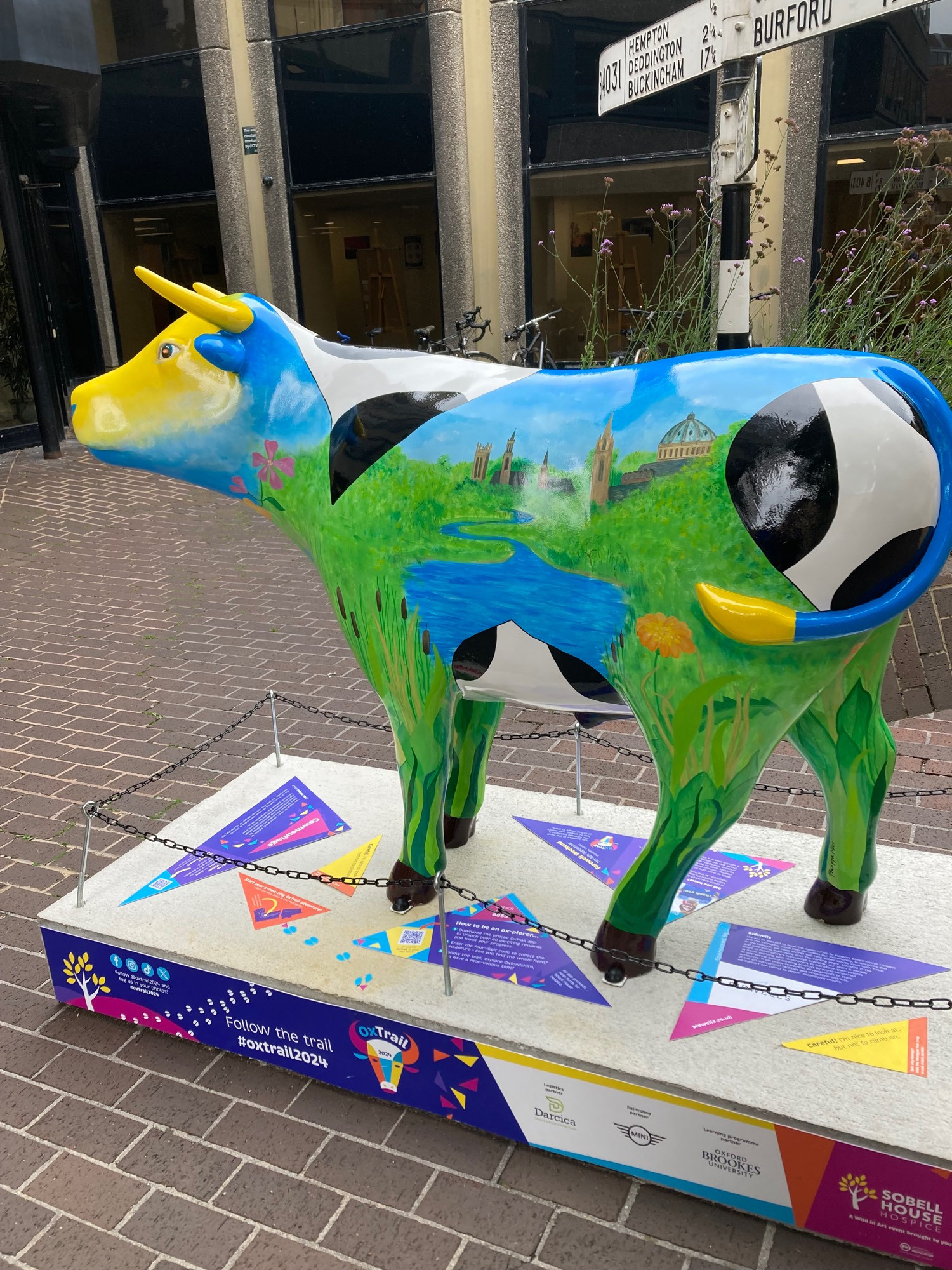 side view of ox sculpture. This side has the black and white cow patches but also green field painted on and water and in the distance oxford buildings