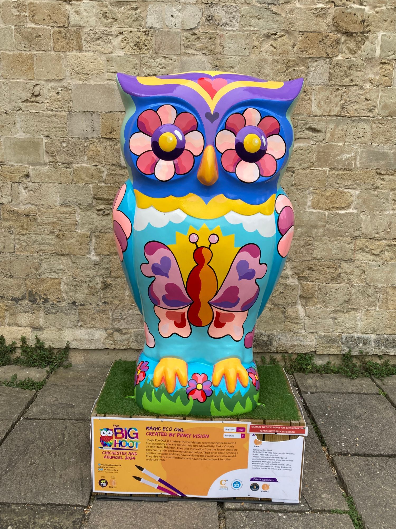 Large owl sculpture, brightly painted with a pink butterfly on its chest, the butterfly has hearts painted on its wings. the owl has flowers for eyes and the main colours are yellow, putple, pink, orange, turquoise and yellow