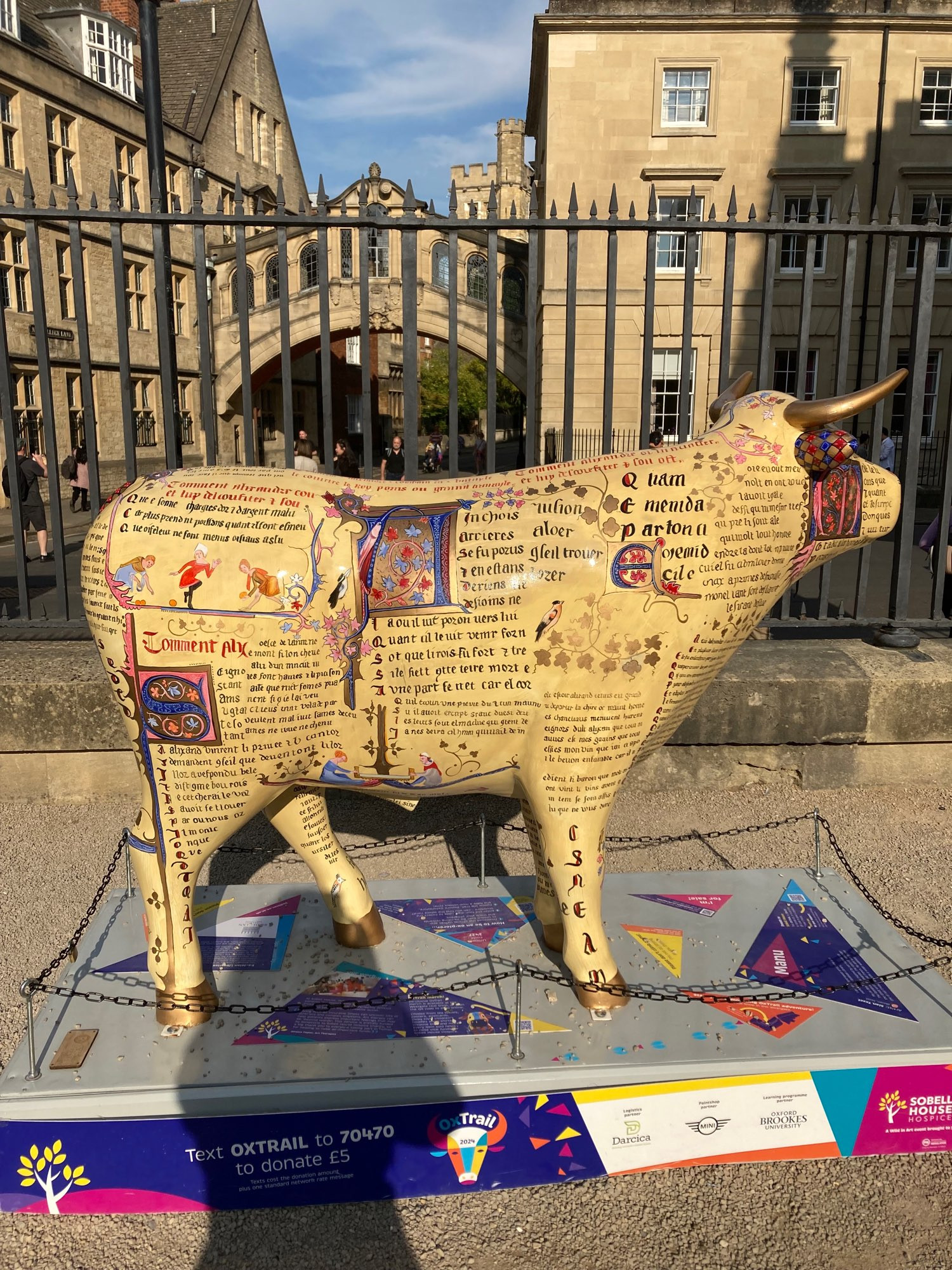 side view of sculpture covered in Latin script and medieval paintings like an ancient manuscript with w walkway between colleges in background