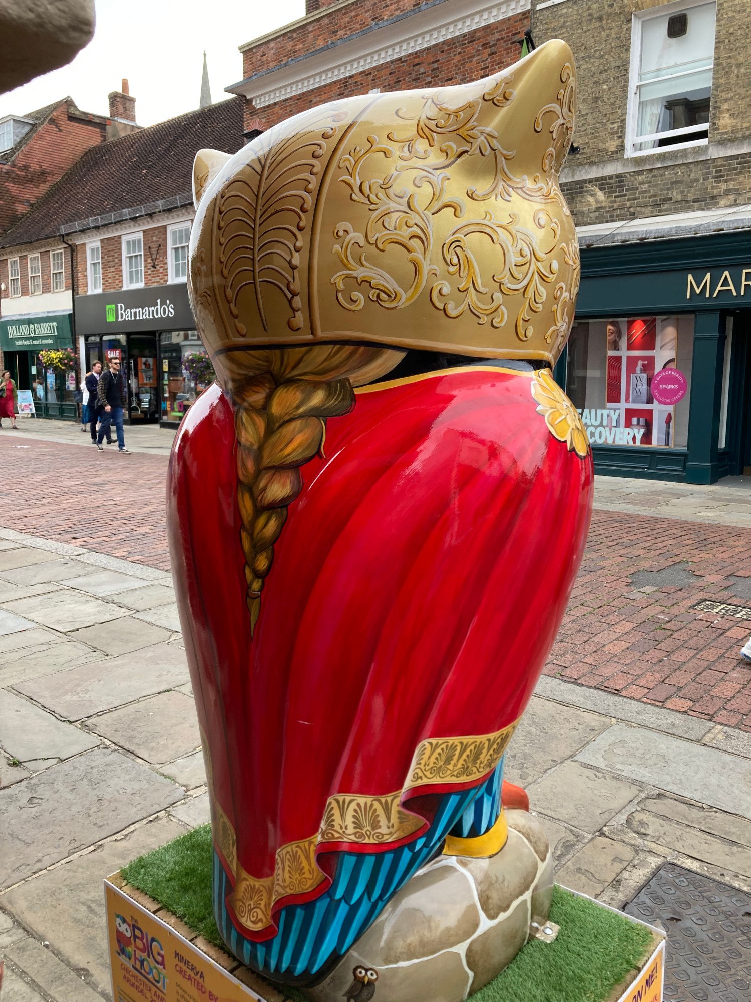 back view of owl sculpture with a gold helmet painted on and plaited blonde hair trailing down a red robe with gold trim