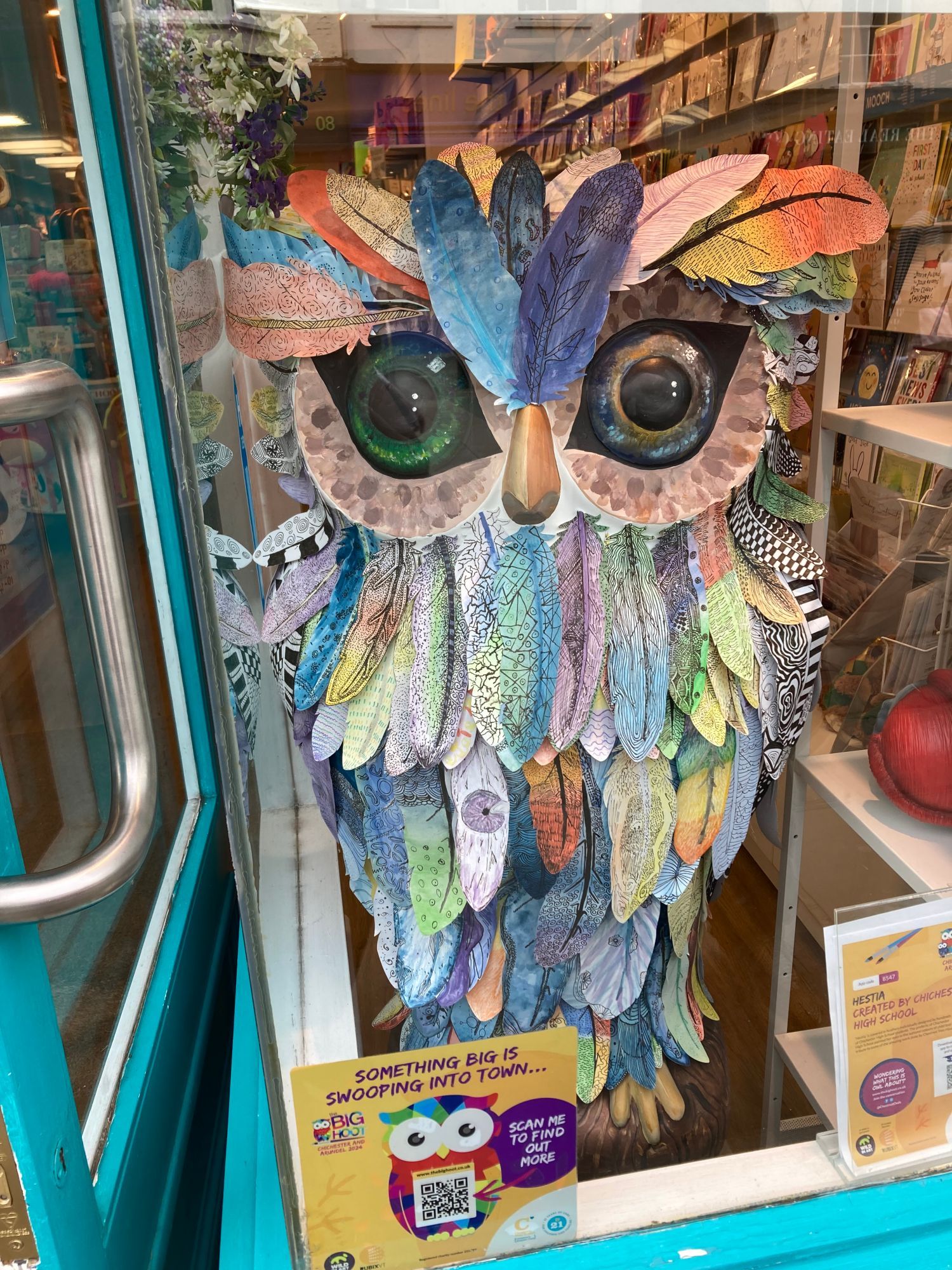 small owl sculpture in charity shop window, it has multiple feathers stuck onto it and painted various pastel colours, it's eyes are large and green. The feathers are in the style of an American Indian headdress.