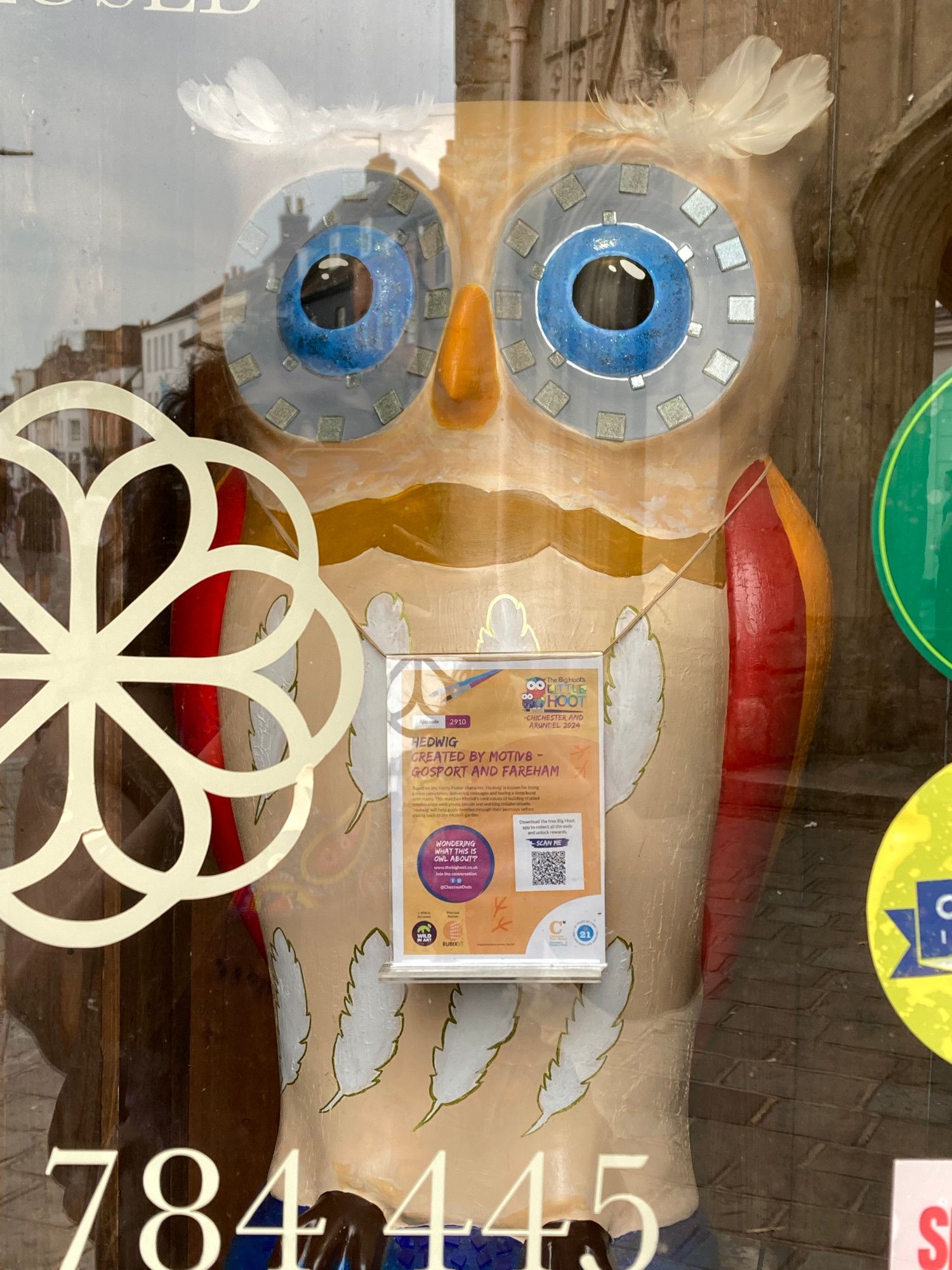 small owl sculpture in shop window, front is pale beige with white feathers painted on, large grey eyes with metallic squares surrounding the blue pupils and real white feathers are stuck on top of its head. The wings are red and orange.