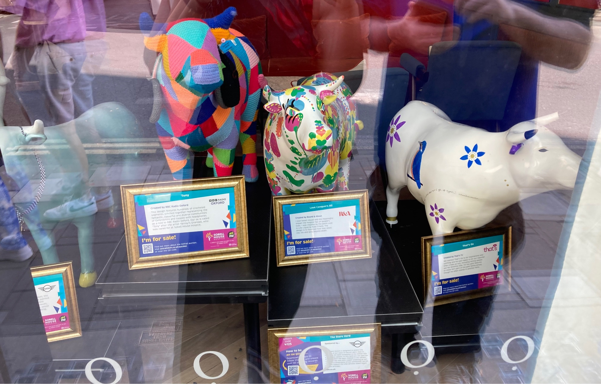 4 mini ox behind glass: pale blue; multicoloured triangles of blue pink orange and green; white with flowers painted in pink blue and yellow; white with blue and purple flowers. The multicoloured one is knitted.