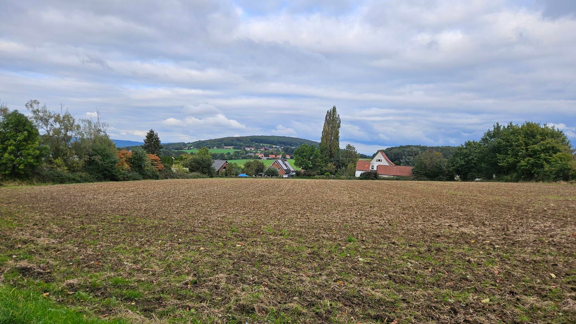 Op de voorgrond een aflopend, bruin en kaal weiland. Halverwege wat bomen eb huizen rechts en links. En in de verte een groen weiland met daarachter een heuvel met wat huisjes en een bos. 