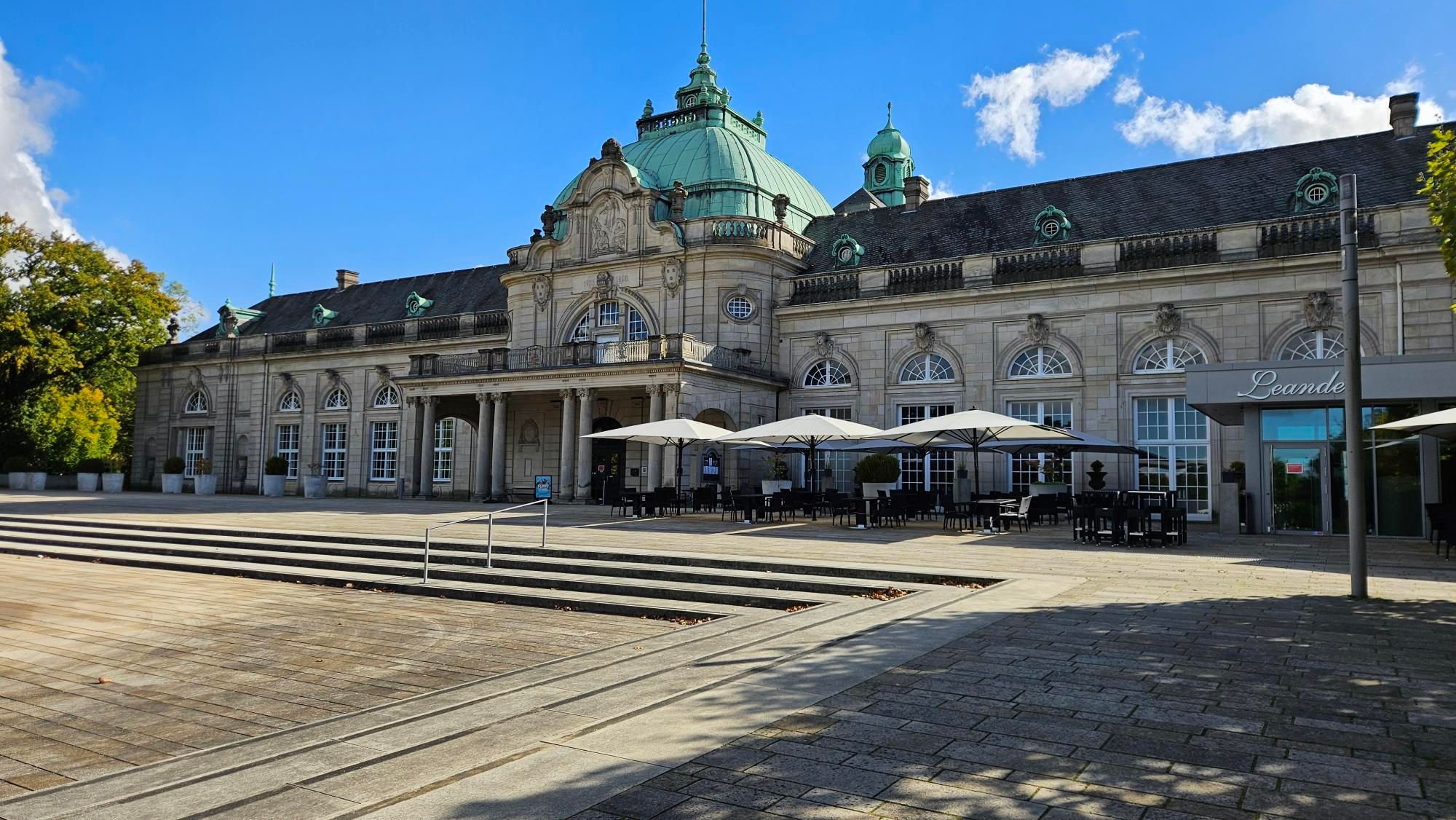 Oud badhuis, tegenwoordig schouwburg, lang gebouw van beige steen met een zwart dak, in het midden onderbroken door een pompeus uitstekend deel met groen dak. Rechtse deel is een restaurant met zitjes ervoor en grote parasols. 