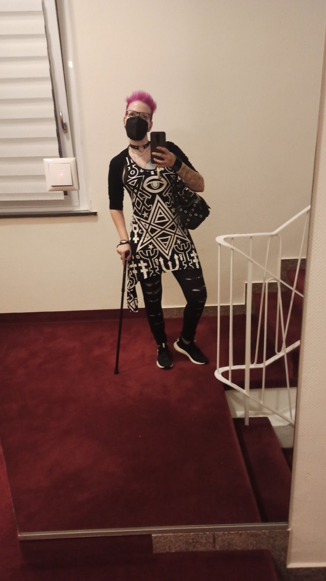 Spiegel Selfie in einem Treppenhaus von Seras, einer weißen, weiblichen Person mit einem kurzen pinken Irokesen. Sie trägt eine Brille, ffp2 Maske, ein schwarz weißes Kleid mit Leggins und Jacke über der Schulterbandage. Sie stützt sich auf ihrem Gehstock ab.