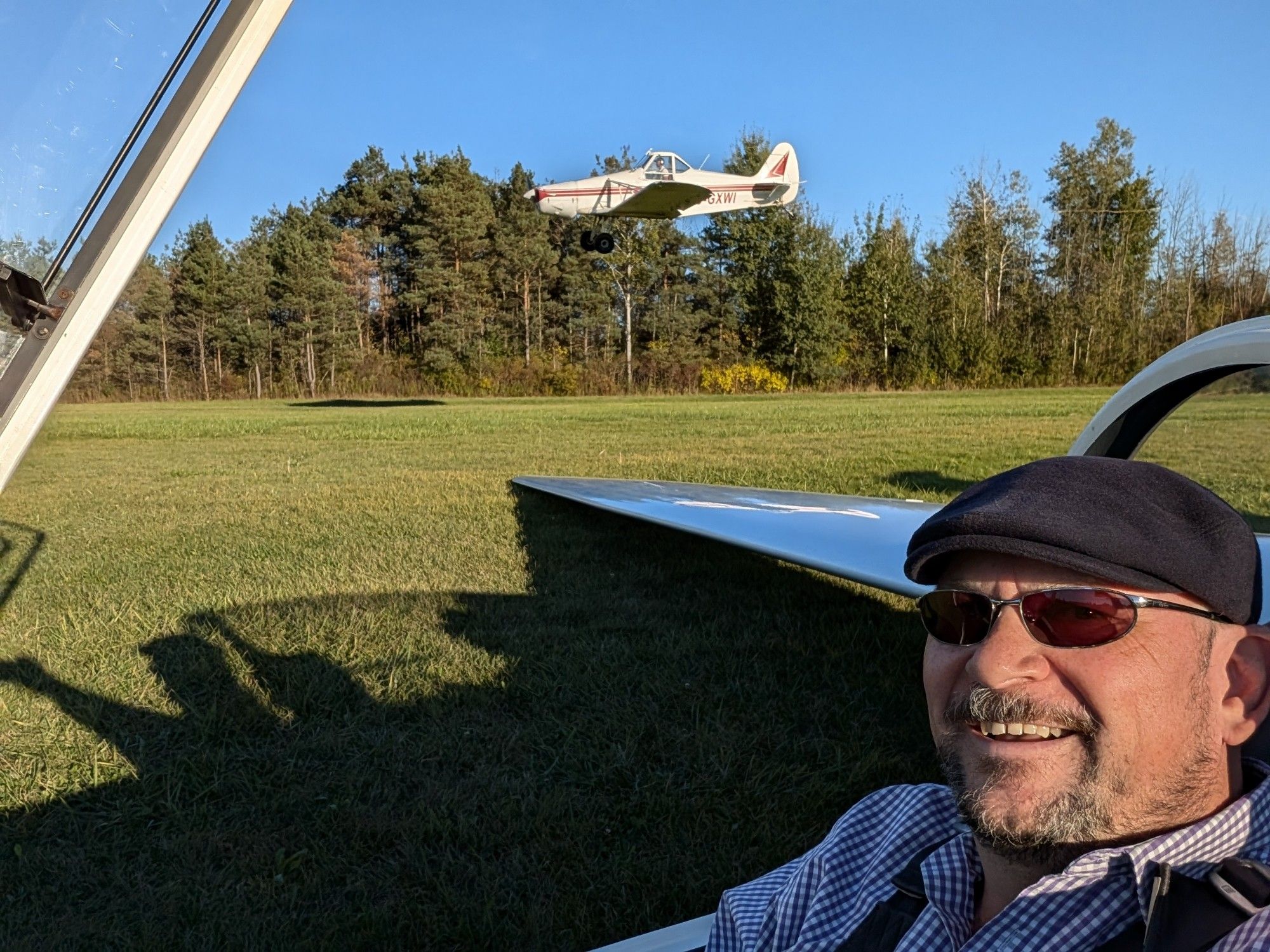 Your handsome pilot narrator in sunglasses and a blue cashmere stetson hat, sitting in a cockpit ready for launch. The right wing stretches to lie on a grass runway as a Piper PA-25 passes in the background while landing.