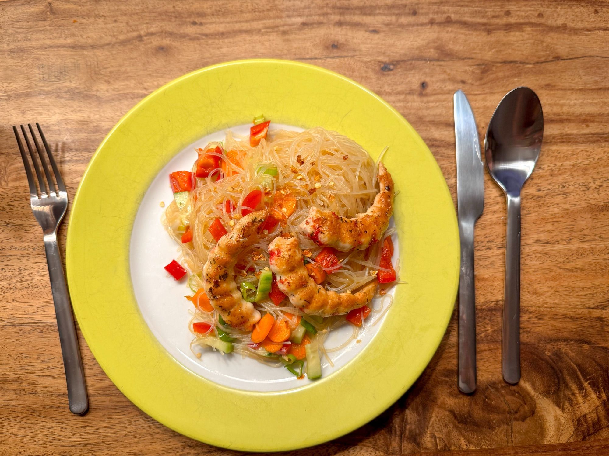 Tisch mit Besteck und grünem Teller. Darauf Glasnudeln dazwischen Paprika, Karotten, Gurken und Frühlingszwiebeln. 
Getoppt mit drei veganen Shrimps und etwas Chiliflocken