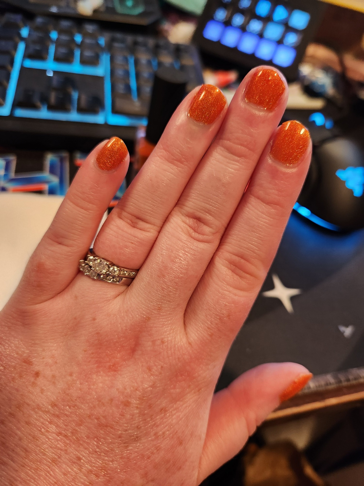 Tap's left hand with her desk in the background. All the nails are sparkling orange and she wears 2 rings on her ring finger.