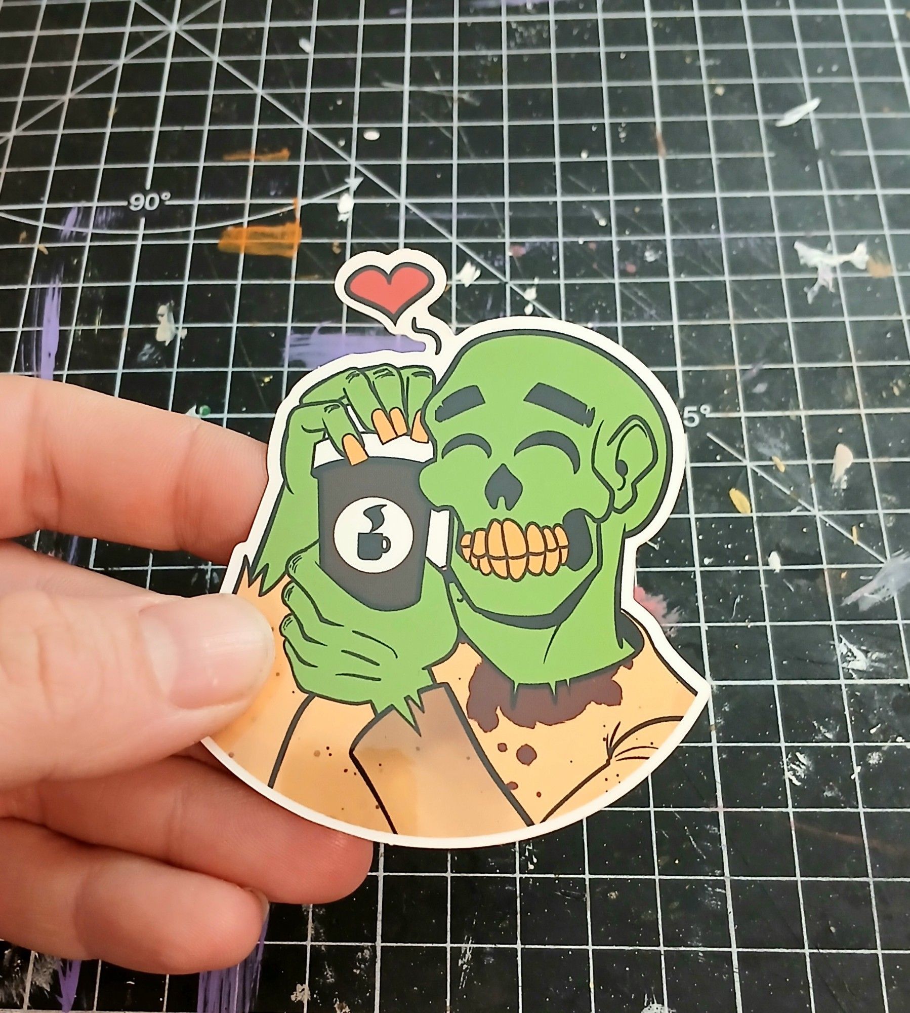 My hand holding a sticker depicting a zombie snuggling a cup of coffee.