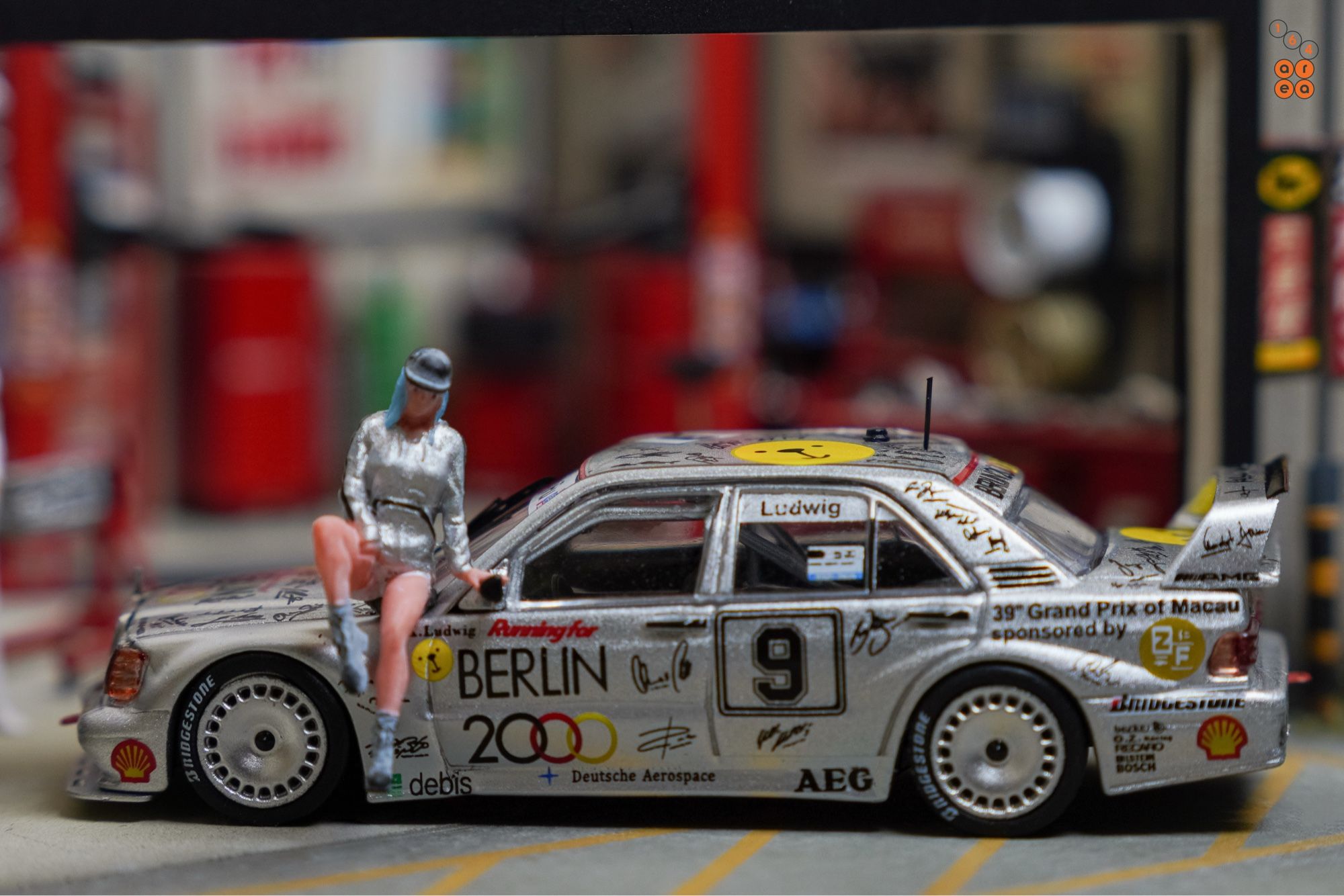 A female in silver suit sit on Mercedes-Benz 190E 2.5-16 Evolution II, Berlin 2000 livery by Tarmac Works in 1:64 scale with 1:64 figure on AREA 164