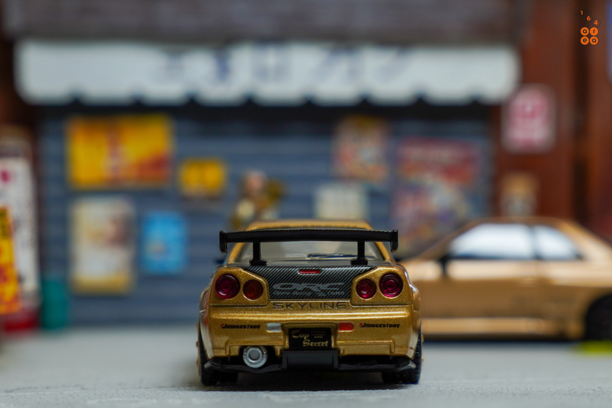 A rear side of Nissan Skyline GT-R (R34) Top Secret, Top Secret Gold by MINI GT in 1:64 scale on AREA 164