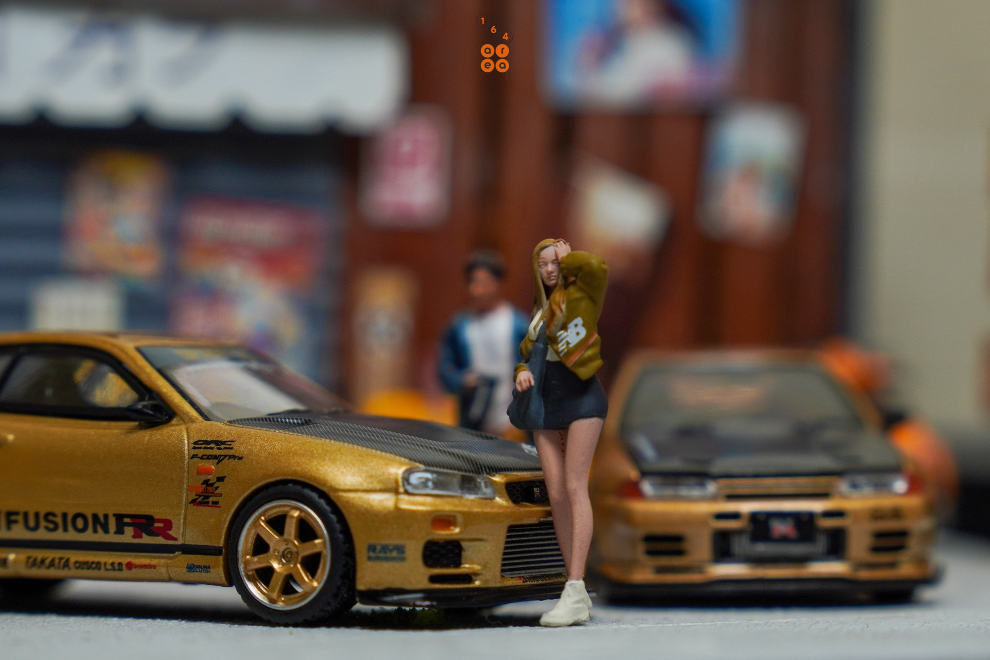 A female stand in front of Nissan Skyline GT-R (R34) Top Secret, Top Secret Gold by MINI GT in 1:64 scale with 1:64 figures on AREA 164