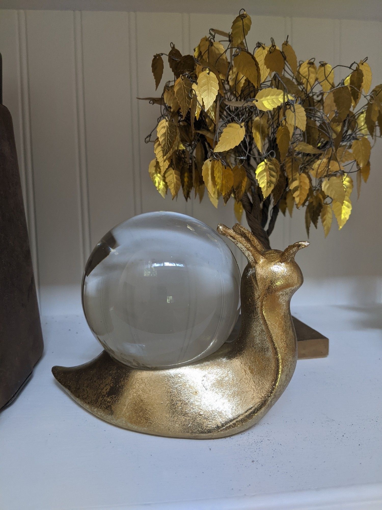 Photo of a bookshelf focused on a gold snail with a crystal ball orb for a shell. In the background there is a golden tree