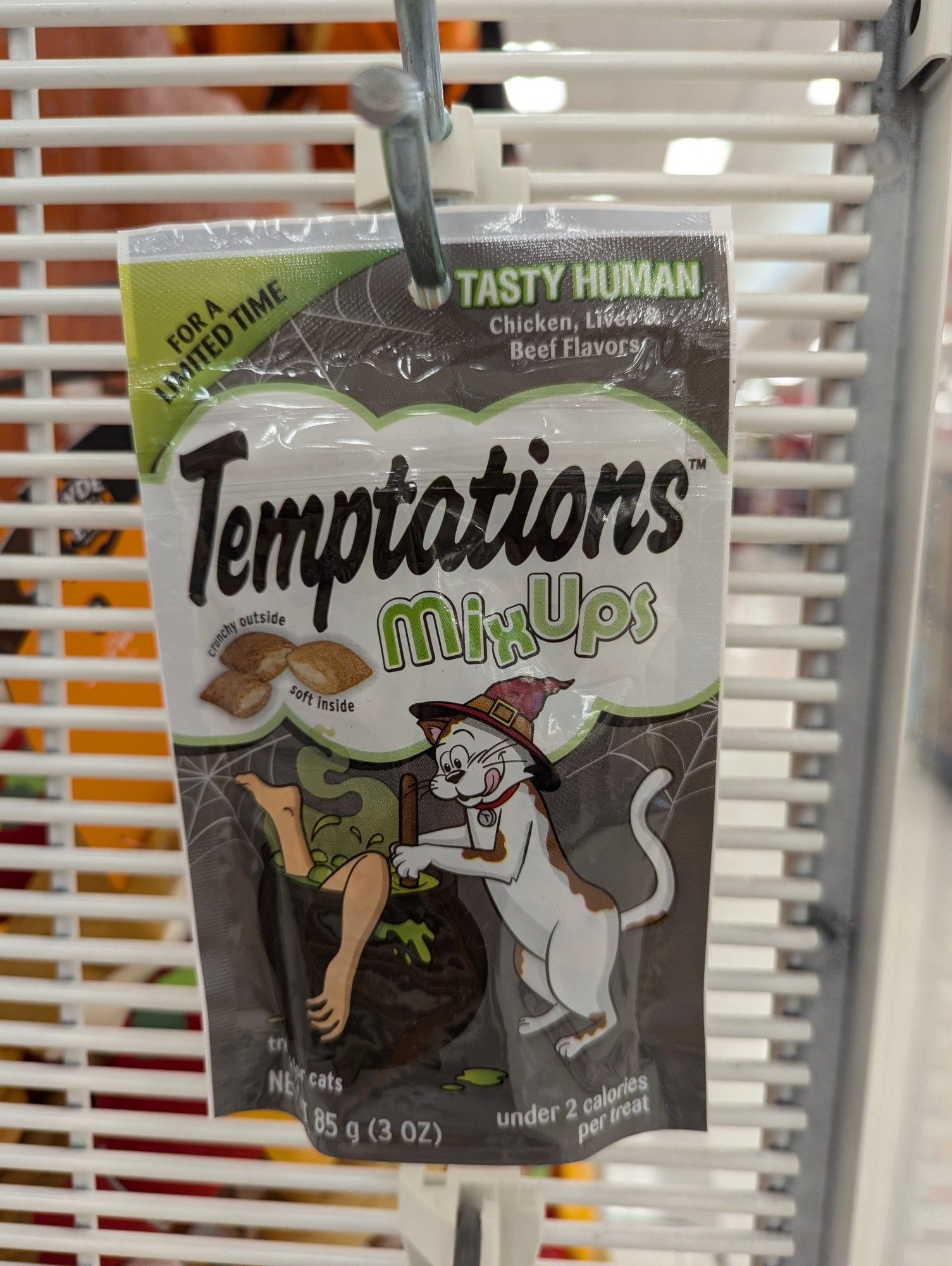 A bag of "Temptations" cat treats in limited edition "Mix Up" Halloween packaging, showing a white cat wearing a witch hat stirring a cauldron full of human limbs. The treats are listed as "Tasty Human" flavor.