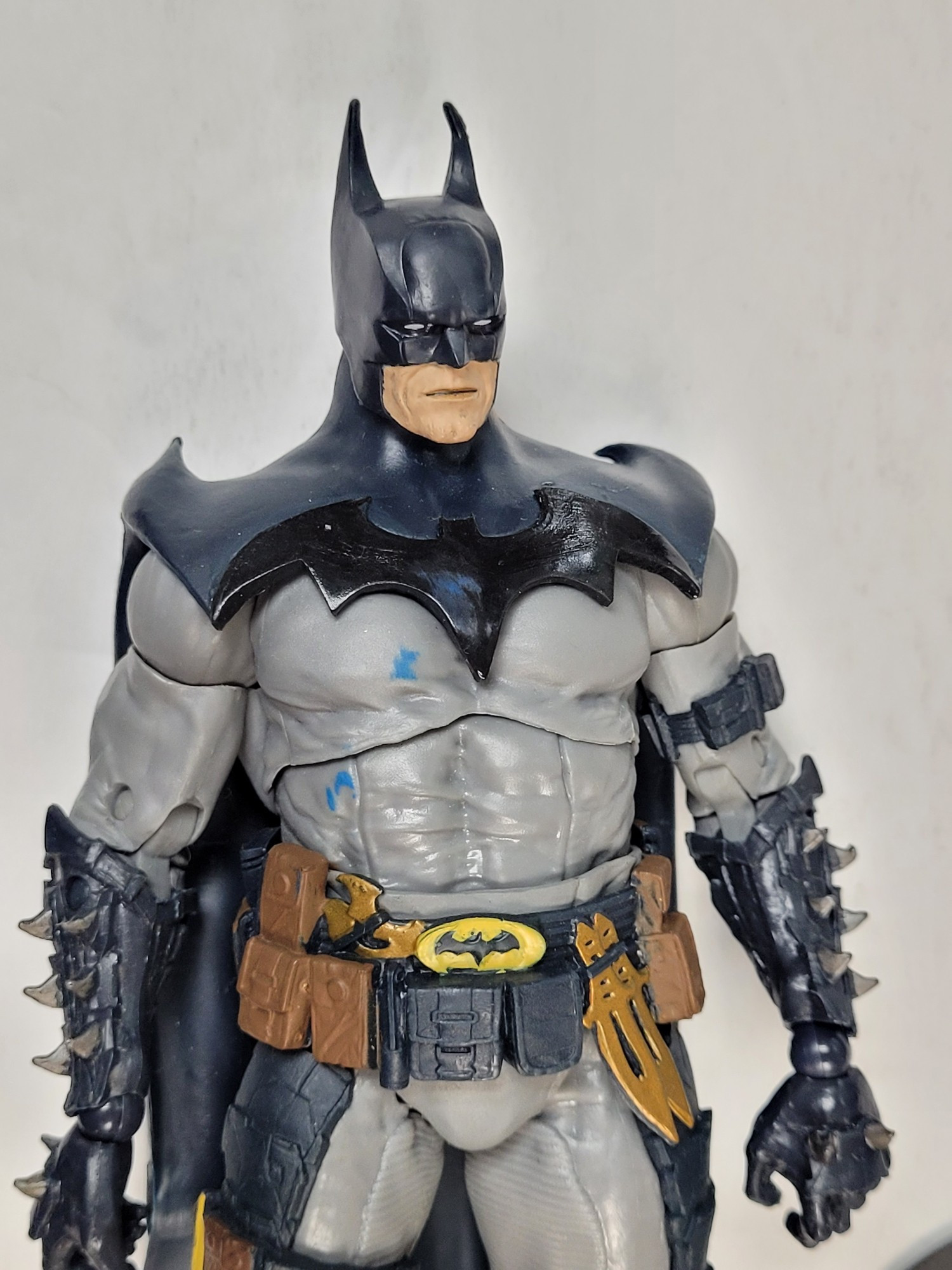 photo of the upper half of todd mcfarlane's action figure of his original design for batman. its the classic blue and grey, with the large black bat emblem linked as part of his shoulder cowl, making it harder to discern from the cape itself. he has spawn style spiked gauntlets, throwing knives on his utility belt, and not pictured are his huge curved knives. this batman will just kill you. he has the yellow oval logo for a belt buckle on his secondary utility belt.