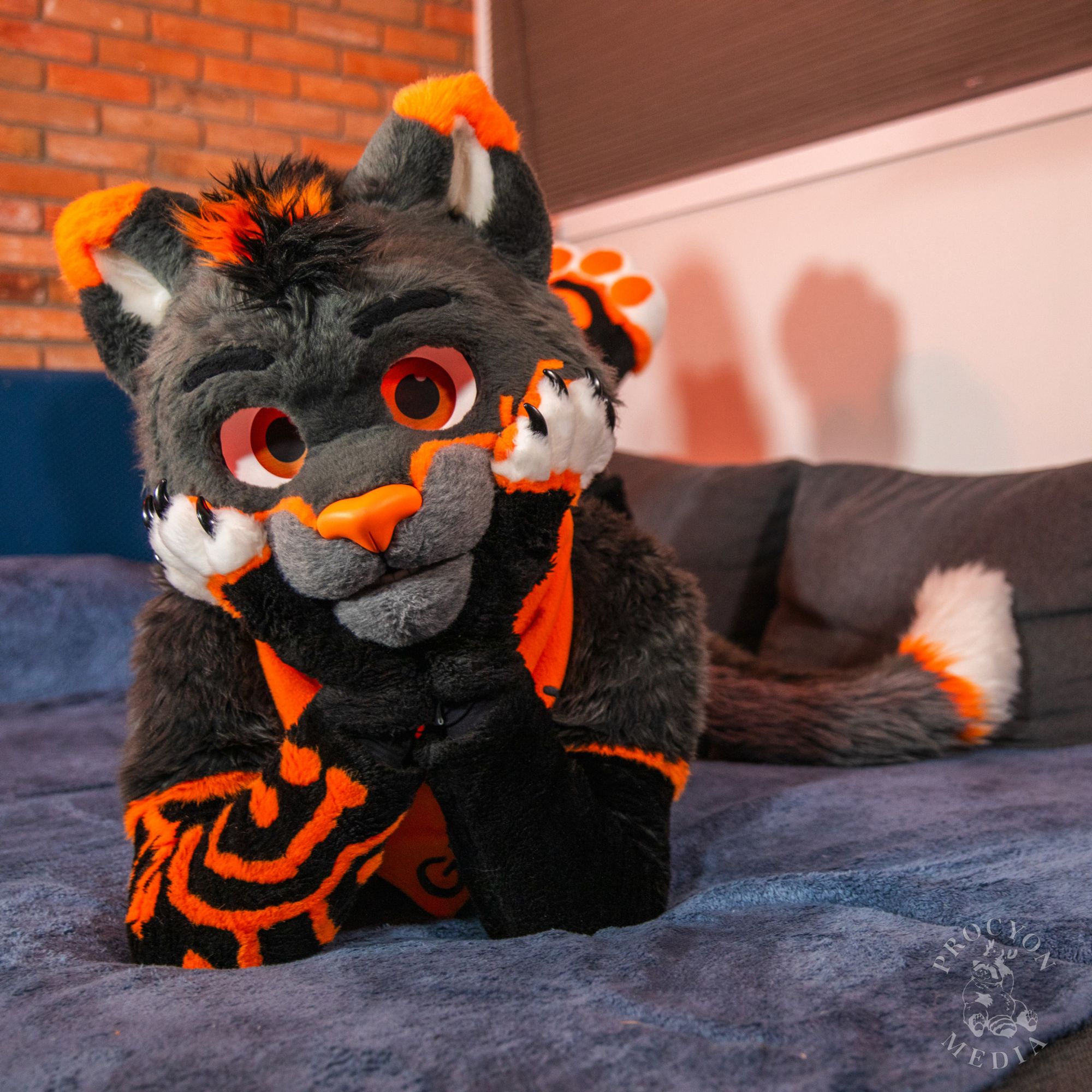 Glitch in fursuit laying belly-side down on a couch/bed, with his head being held up by resting his chin/muzzle on his hands and his arms making a triangle shape. His head is tilted slightly as his legs kick idly in the background.