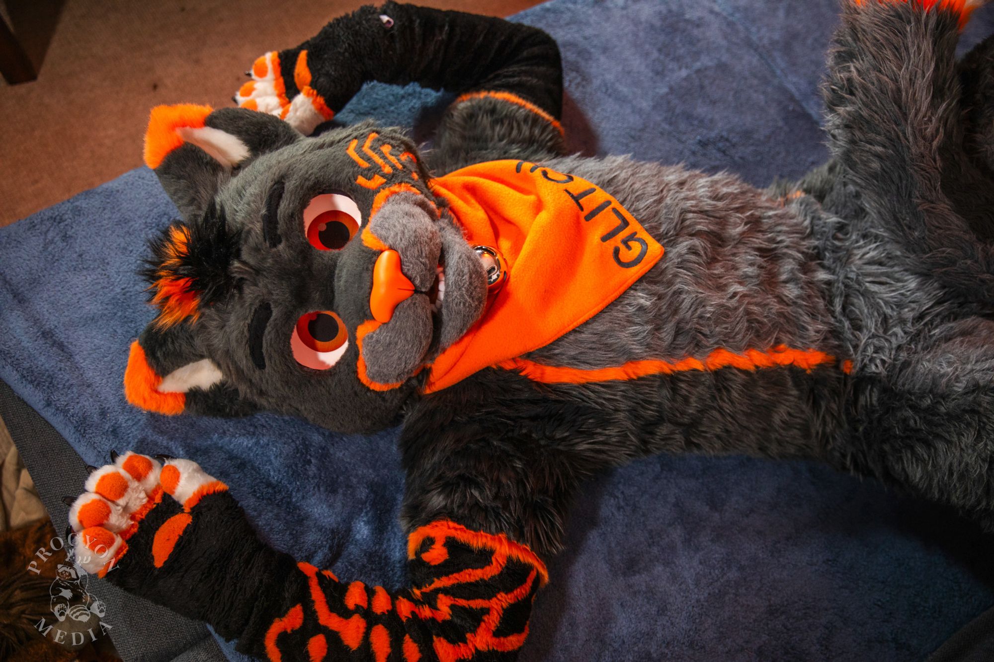 Glitch in fursuit laying face up on a bed, with his arms up and palms showing, resting on each side of his head. The camera is positioned above him, looking straight down. He is wearing an orange bandana around his neck that says "Glitch" and his jaw is slightly open, showing his bottom row of teeth.