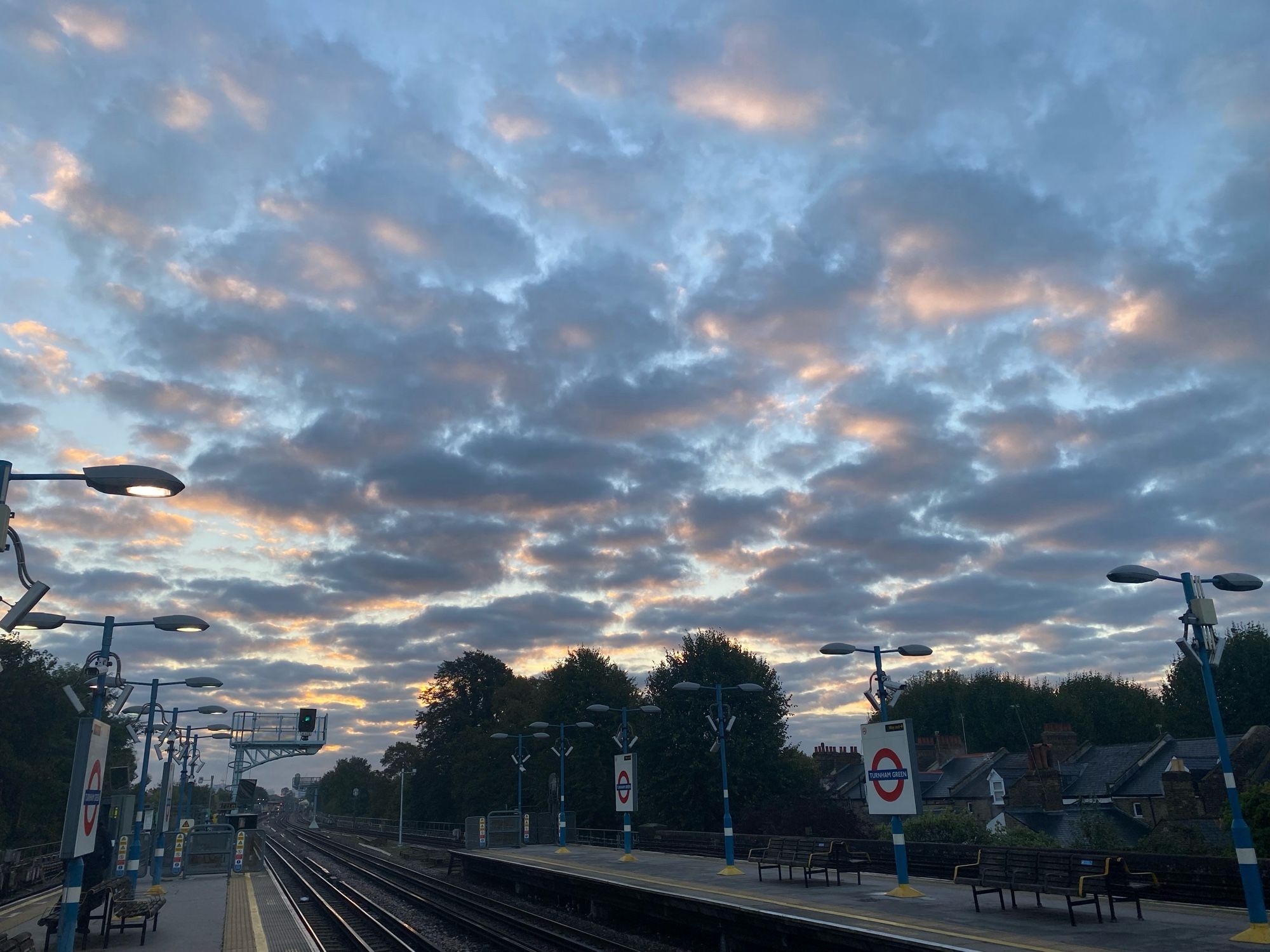 Train platform and tracks with wide sky above showing dotted violet, goldish pink and blue clouds in a jigsaw or mosaic pattern. There is a tube sign which says Turnham Green.