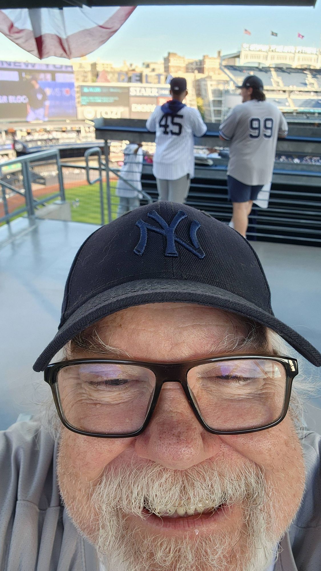 selfie of an old white man with eyeglasses and a white beard, wearing a black on black Yankees cap, with the interior of Yankees Stadium in the background. He's grinning!