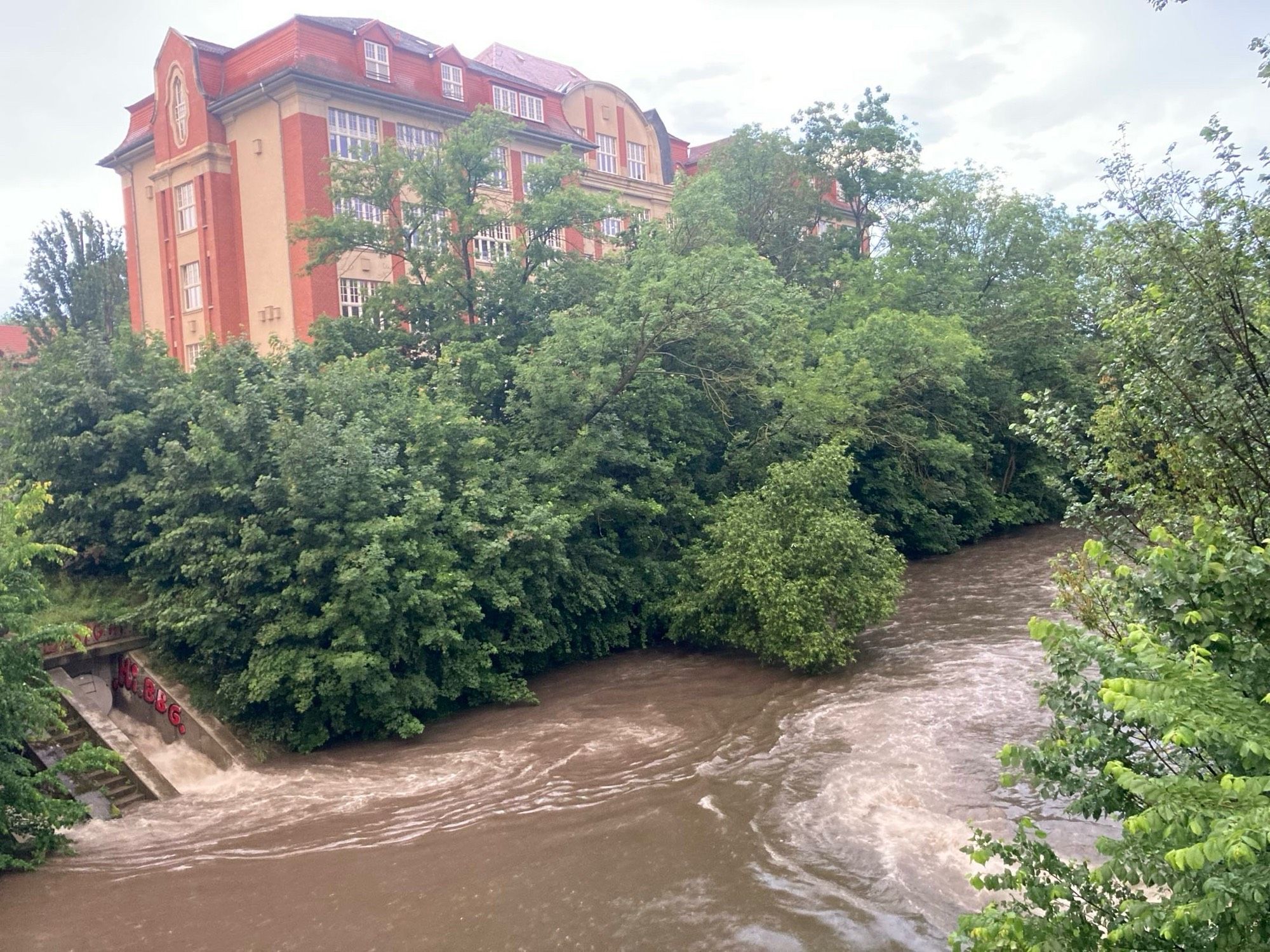 Überlauf am Regenrückhaltebecken entlässt Wasser aus Kanalisation in den Fluss, dahinter Uferbäume und eine Schule