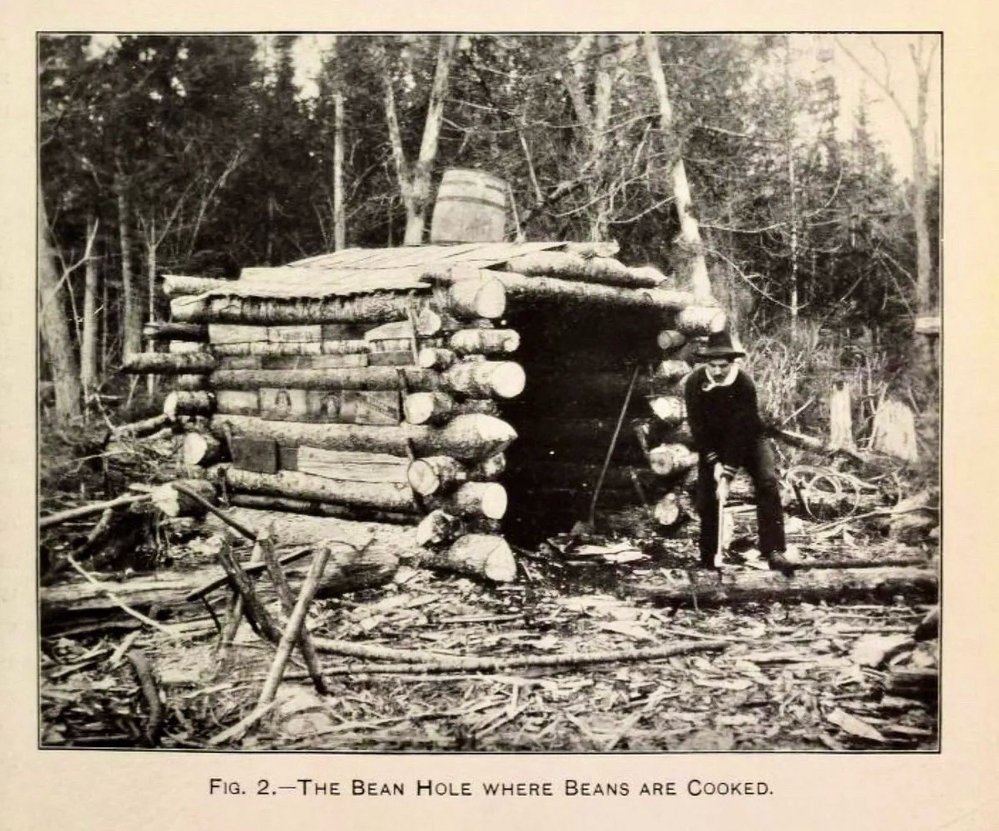 b&w photo of a 19th century cabin with a man sitting outside. the caption says “the bean hole where beans are cooked”