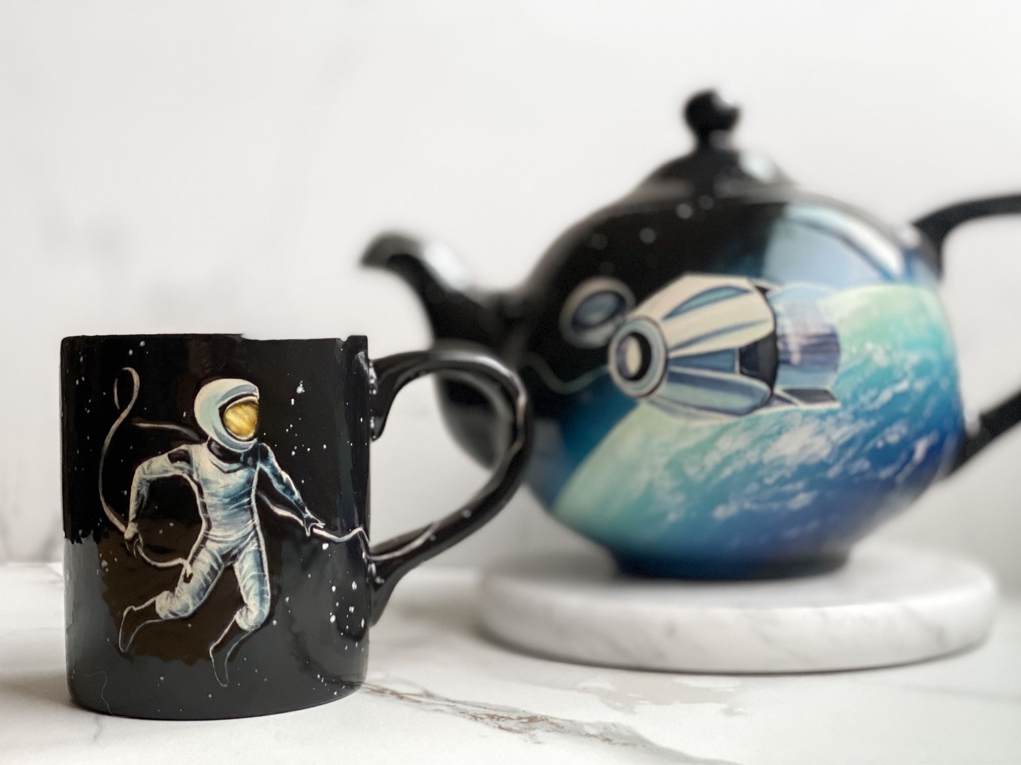 Teapot featuring capsule orbiting earth, mug featuring astronaut connected to tether leading back to capsule on teapot.