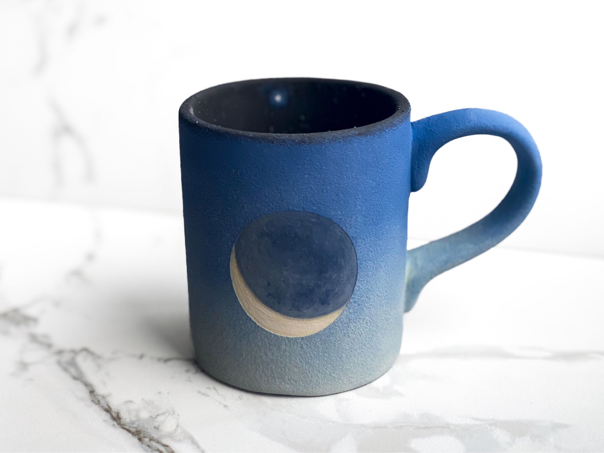 Mug that features crescent moon against a deep blue sky while the inside is black with tiny Venus at the top toward the rim.