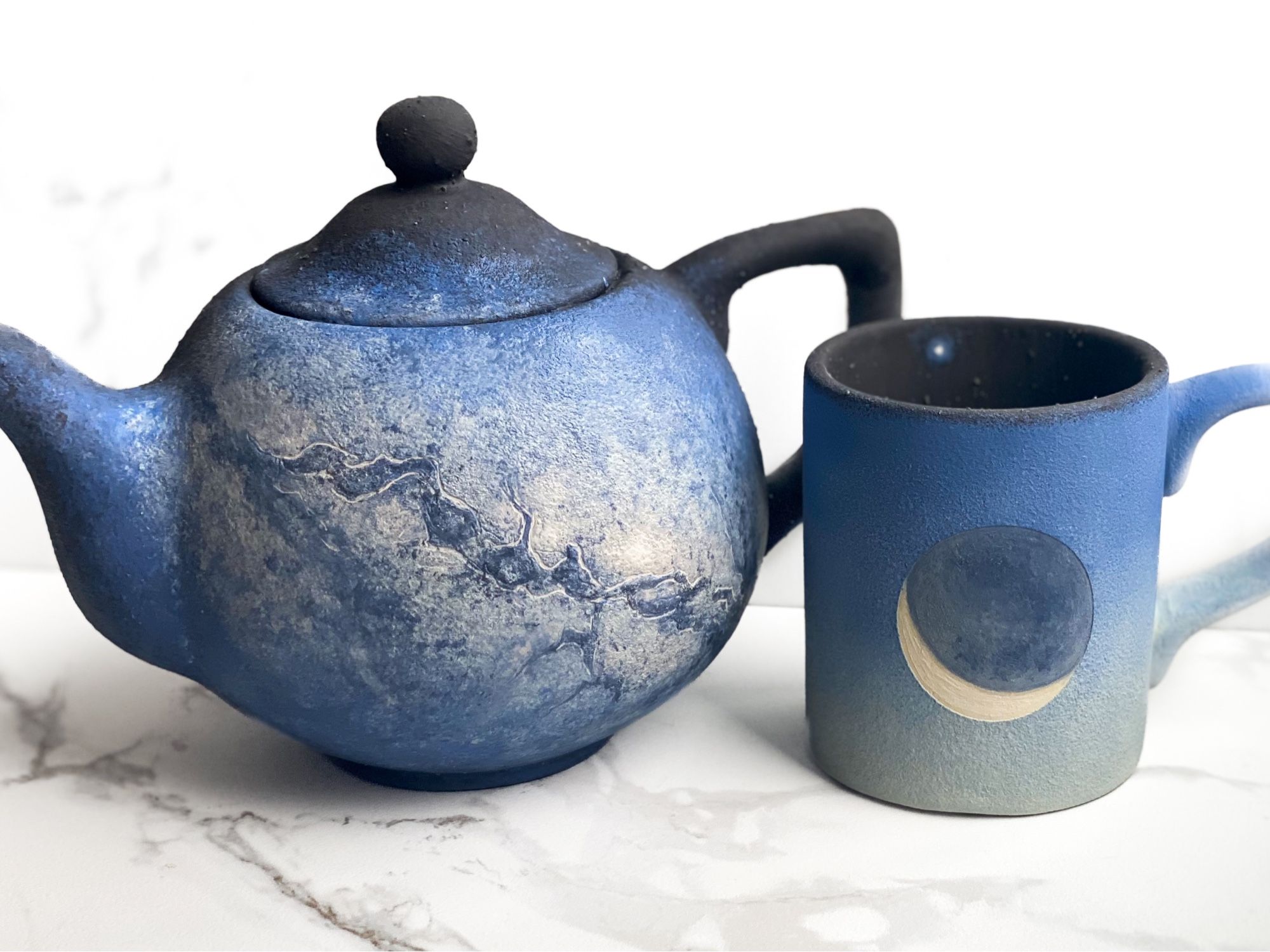 Teapot and mug. Teapot features soft view of Milky Way galaxy and mug features crescent moon against a deep blue sky while the inside is black with tiny Venus at the top toward the rim.