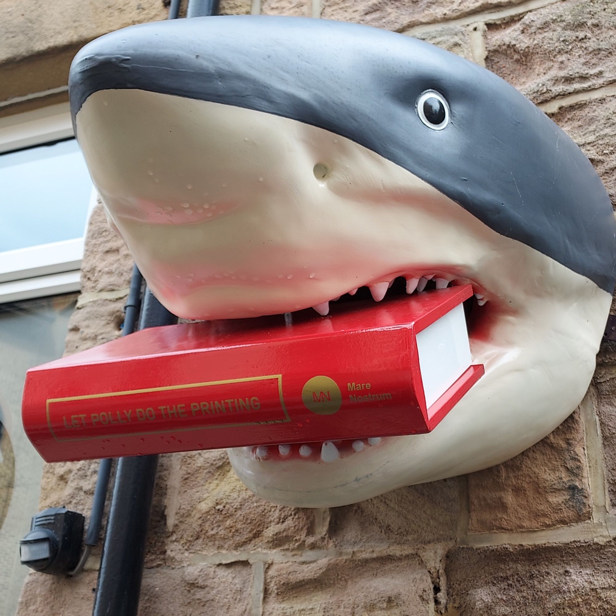 Giant shark emerging from the Mare Nostrum Group office building holding a book titled 'Let Polly Do The Printing'