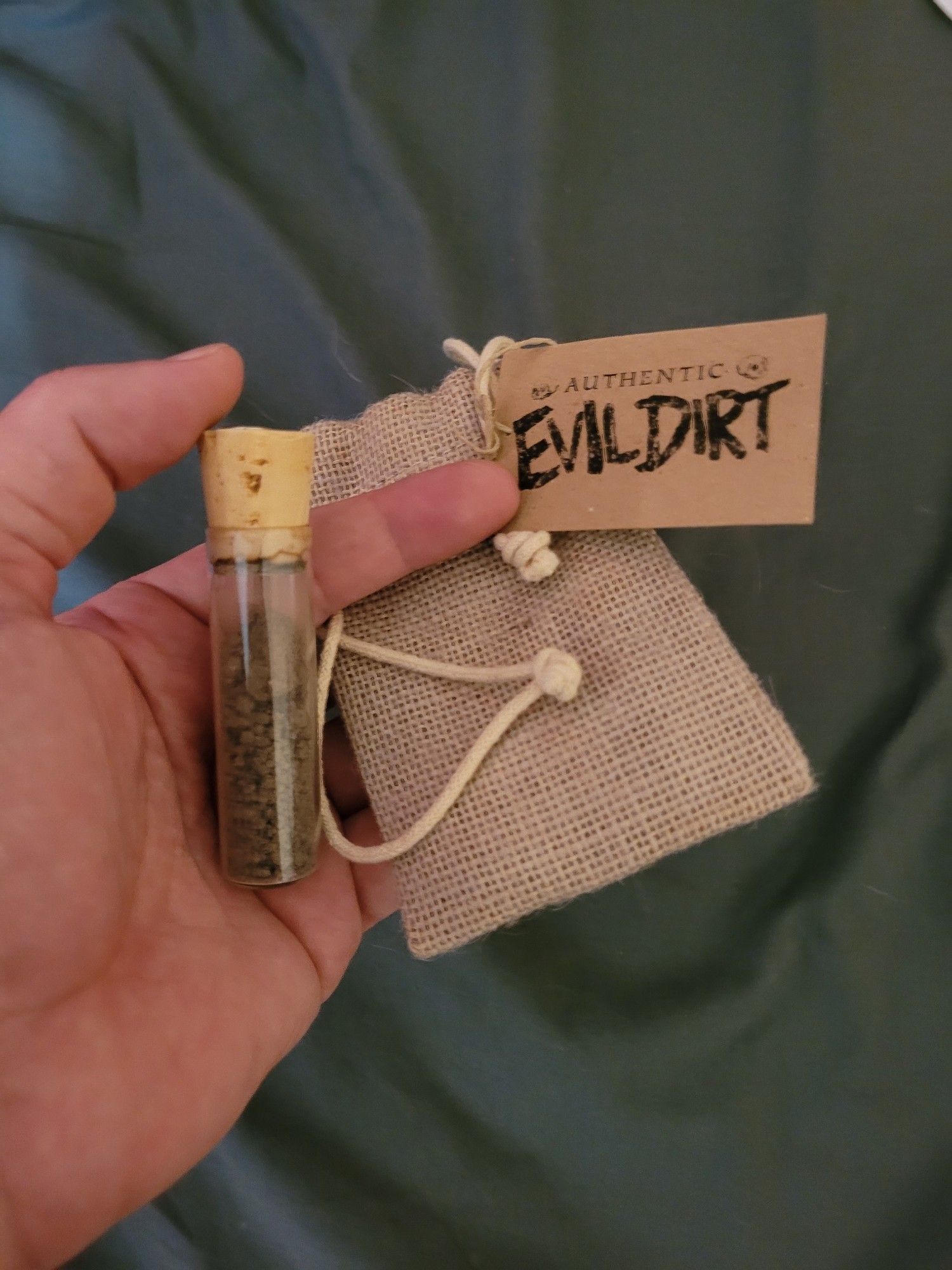 A picture of a human hand holding a clear vial containing a small amount of dirt, along with a card with the words "Evil Dirt" printed in a creepy font.